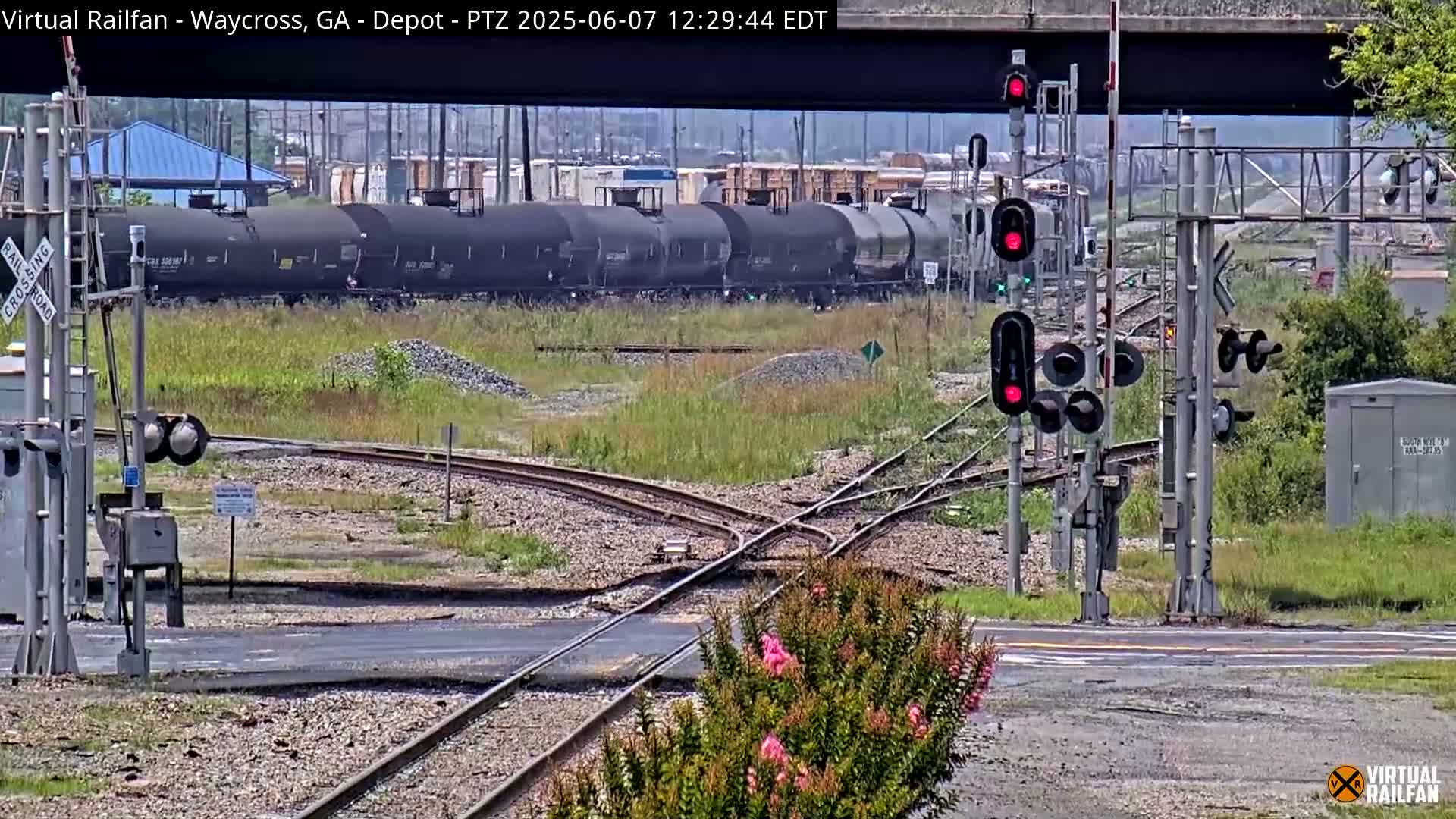 A train of tanker cars sits near a railway crossing with active traffic signals under a partly sunny sky.