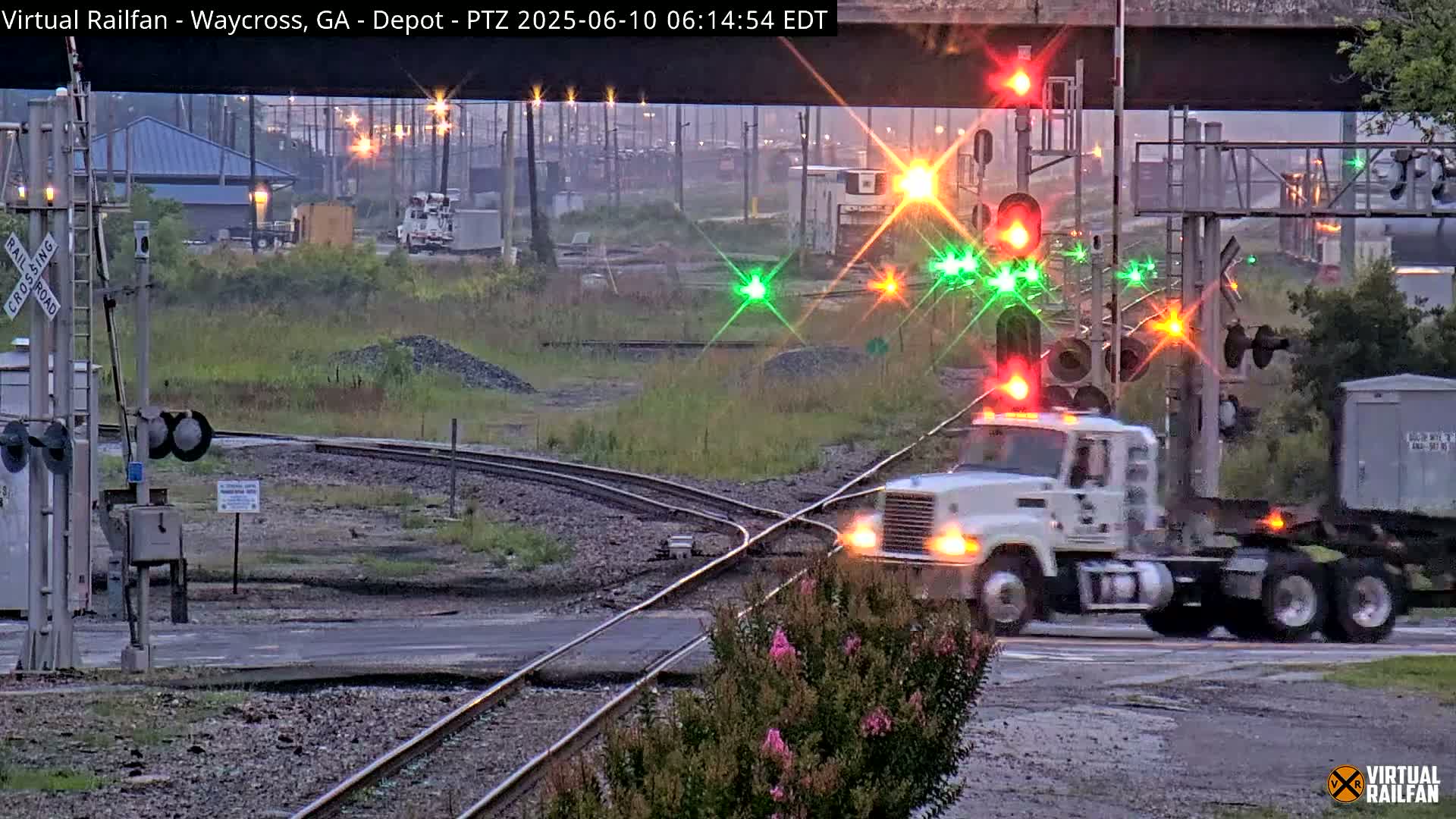 A white semi-truck is crossing a railroad intersection at dawn under a mostly clear sky, with railroad signals flashing red and green.