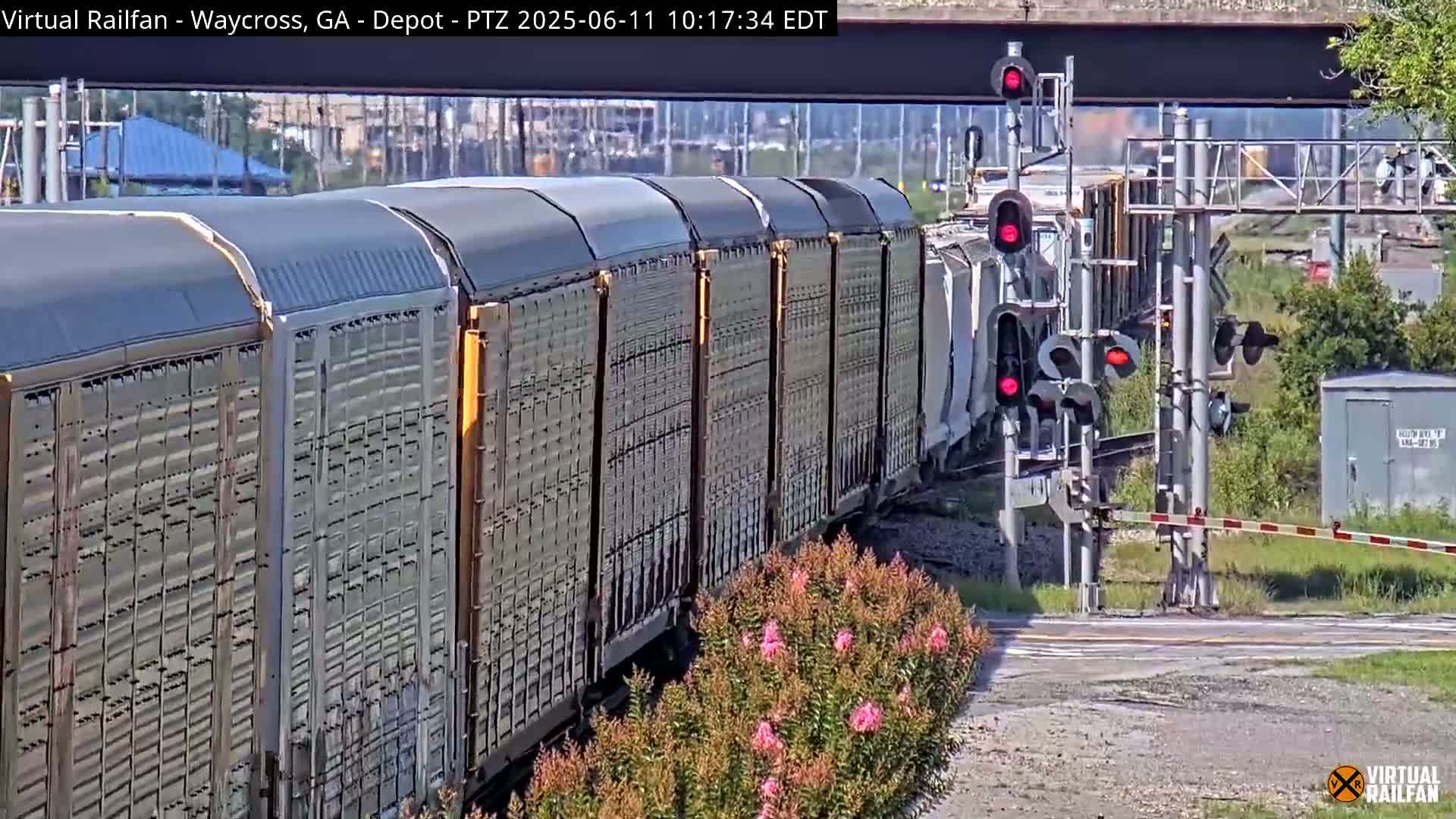 A long freight train passes a railroad crossing under a bridge on a sunny day.