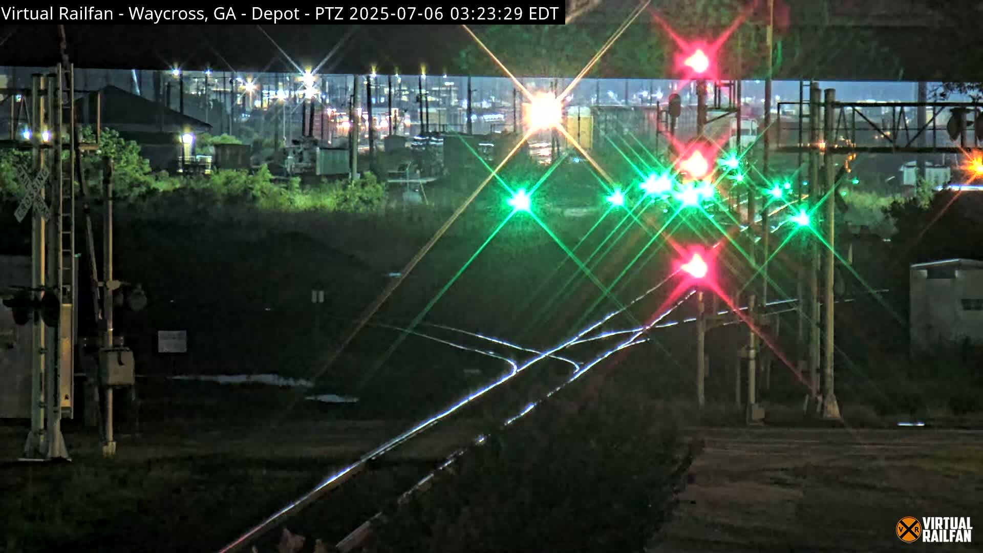 At night, a railroad crossing with illuminated green and red signal lights is shown, with an industrial area visible in the background.