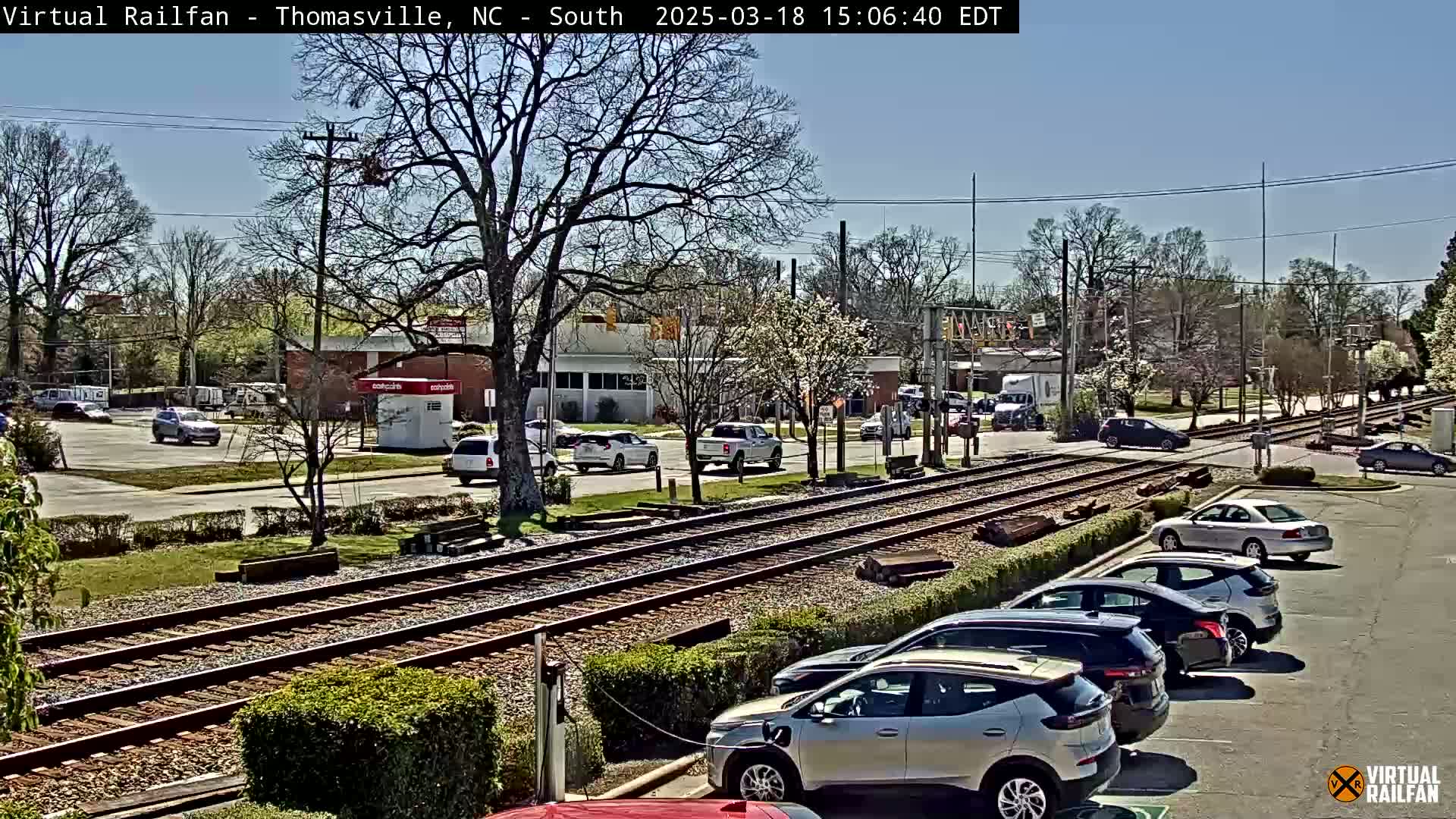 A sunny day shows a railroad crossing with several cars parked alongside and driving on a street near a few buildings, surrounded by leafed trees.