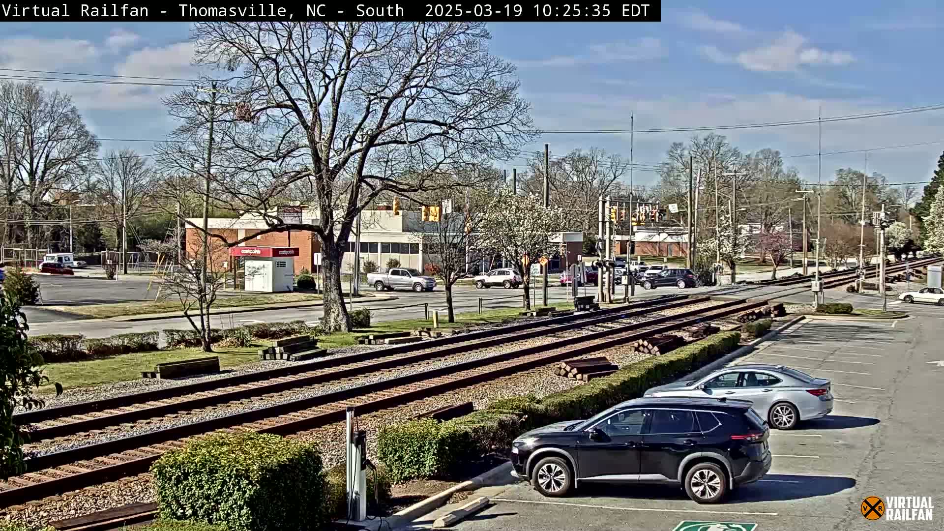 A sunny day shows railroad tracks crossing a road in a town with several cars parked and driving, and trees beginning to bud.