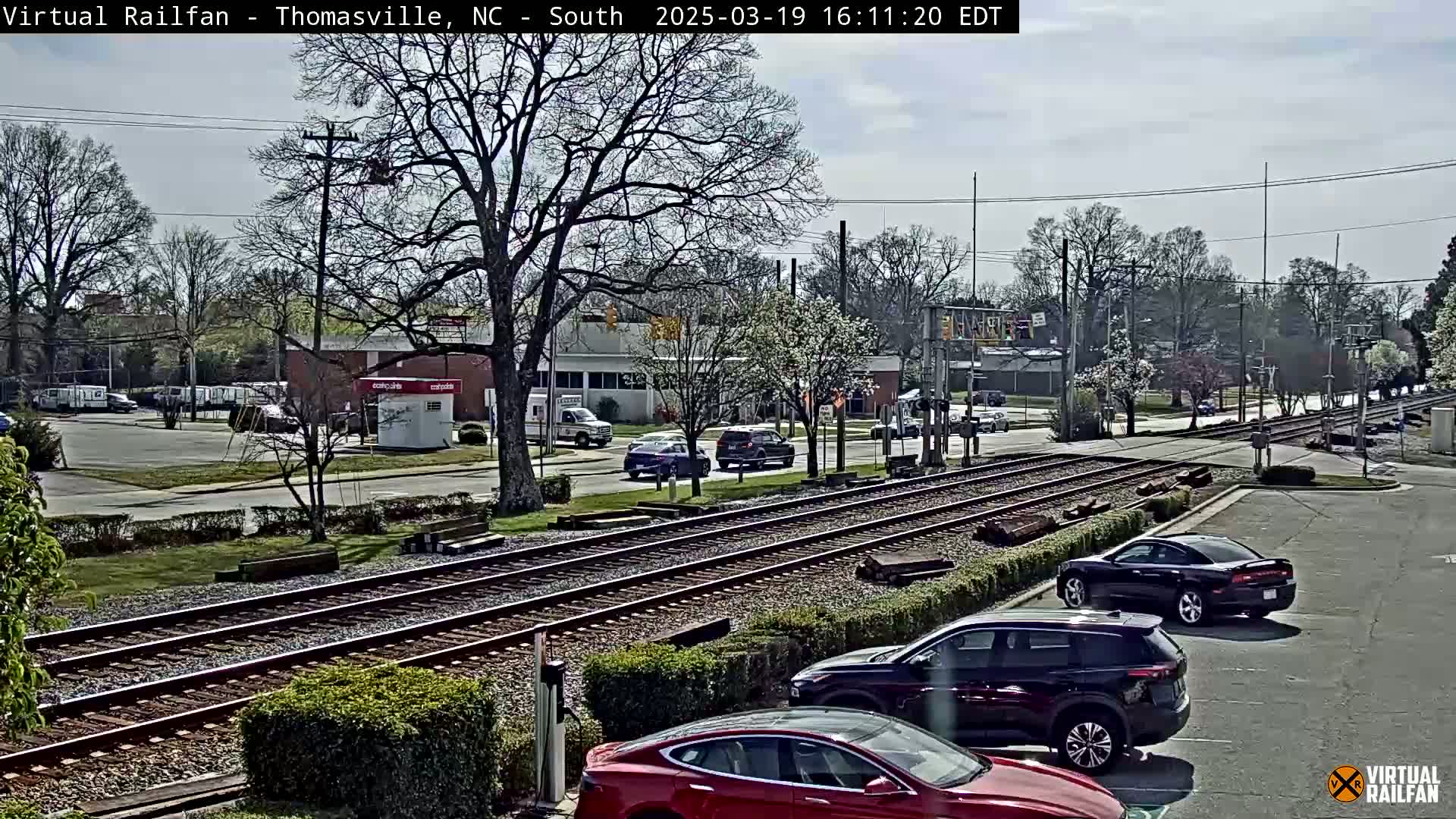 A partly cloudy day shows a view of railroad tracks intersecting a road with several cars parked and driving, passing by low-lying buildings and trees.
