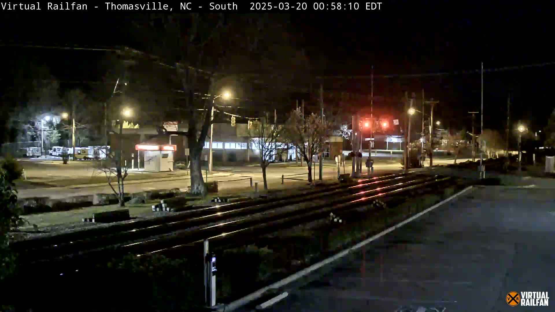 At night, a view of railroad tracks crosses a street with streetlights illuminating a mostly empty intersection and a few parked vehicles.