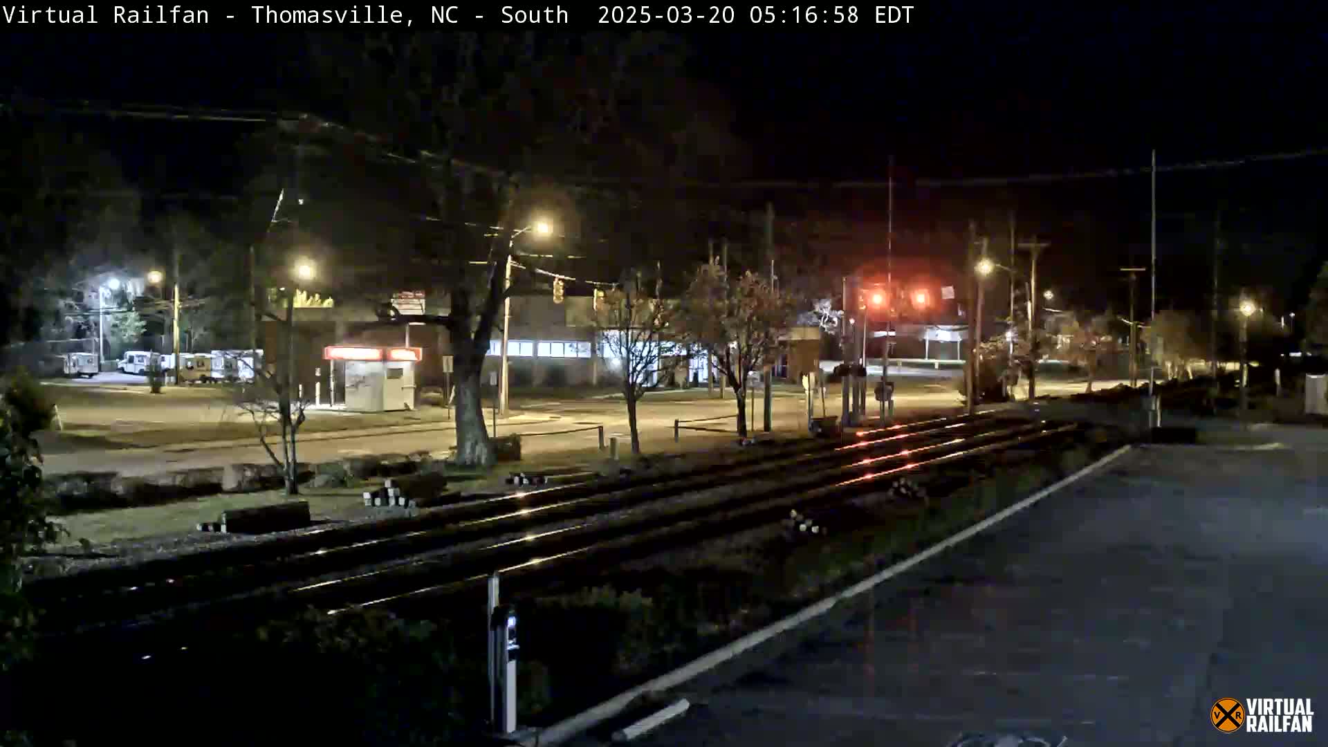 At night, railroad tracks run through a dimly lit town, with streetlights illuminating a road and parked vehicles.