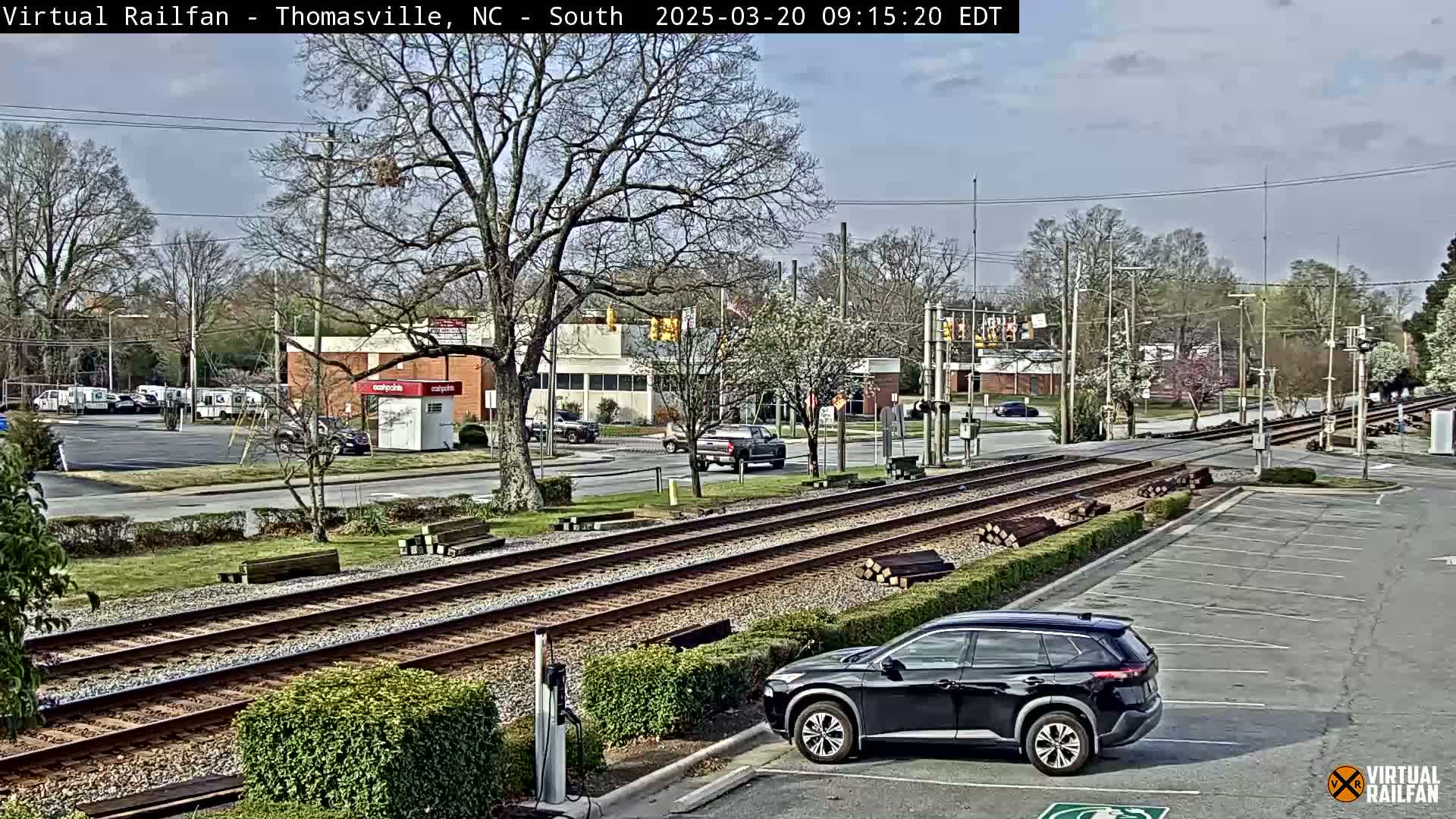 A partly sunny day shows a view of railroad tracks crossing a street, with several vehicles in a parking lot and on the road, surrounded by trees and shrubs.