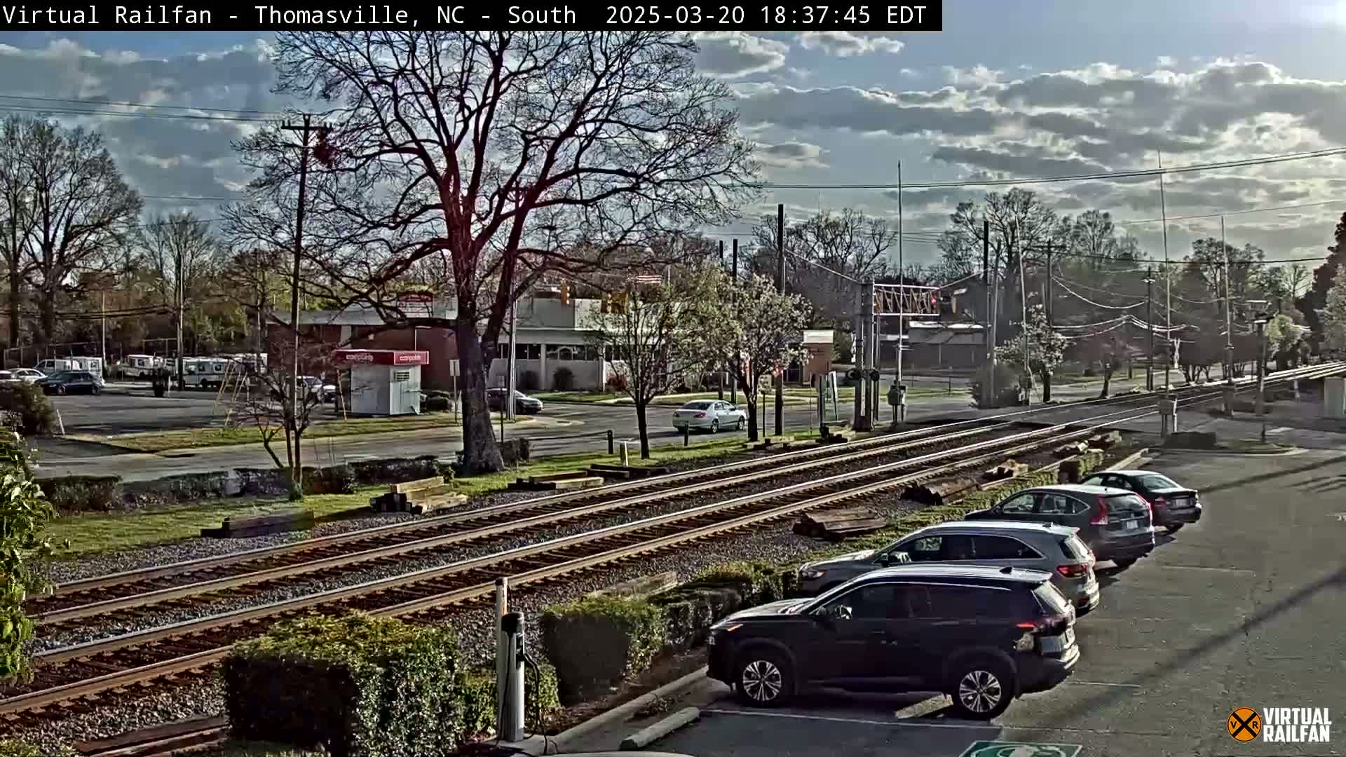 A partly cloudy daytime scene shows several cars parked near railroad tracks that run through a town with buildings and trees.
