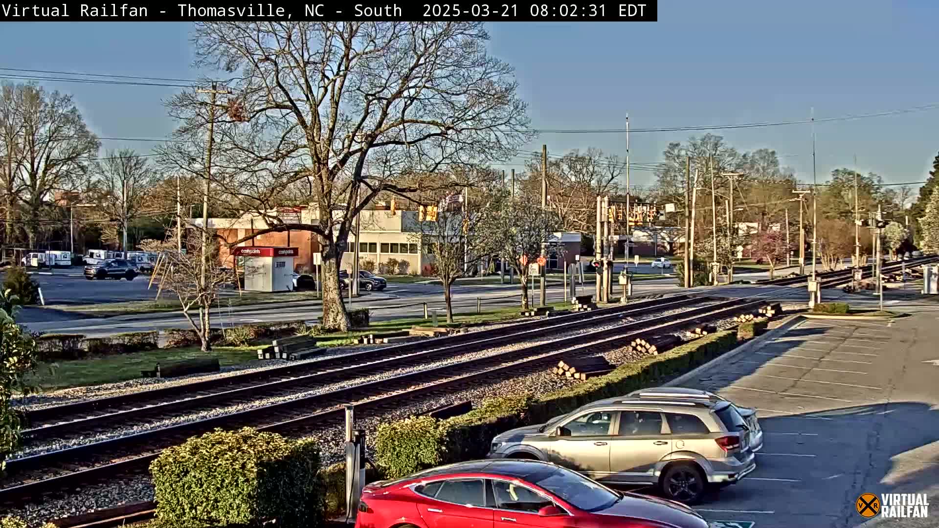 A sunny day shows a view of railroad tracks intersecting a road, with several cars parked nearby in a parking lot, and a few cars driving on the road.