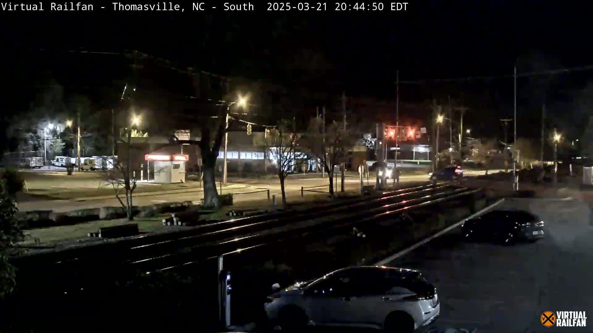 A nighttime view of a railroad crossing with several cars parked nearby, under clear skies.