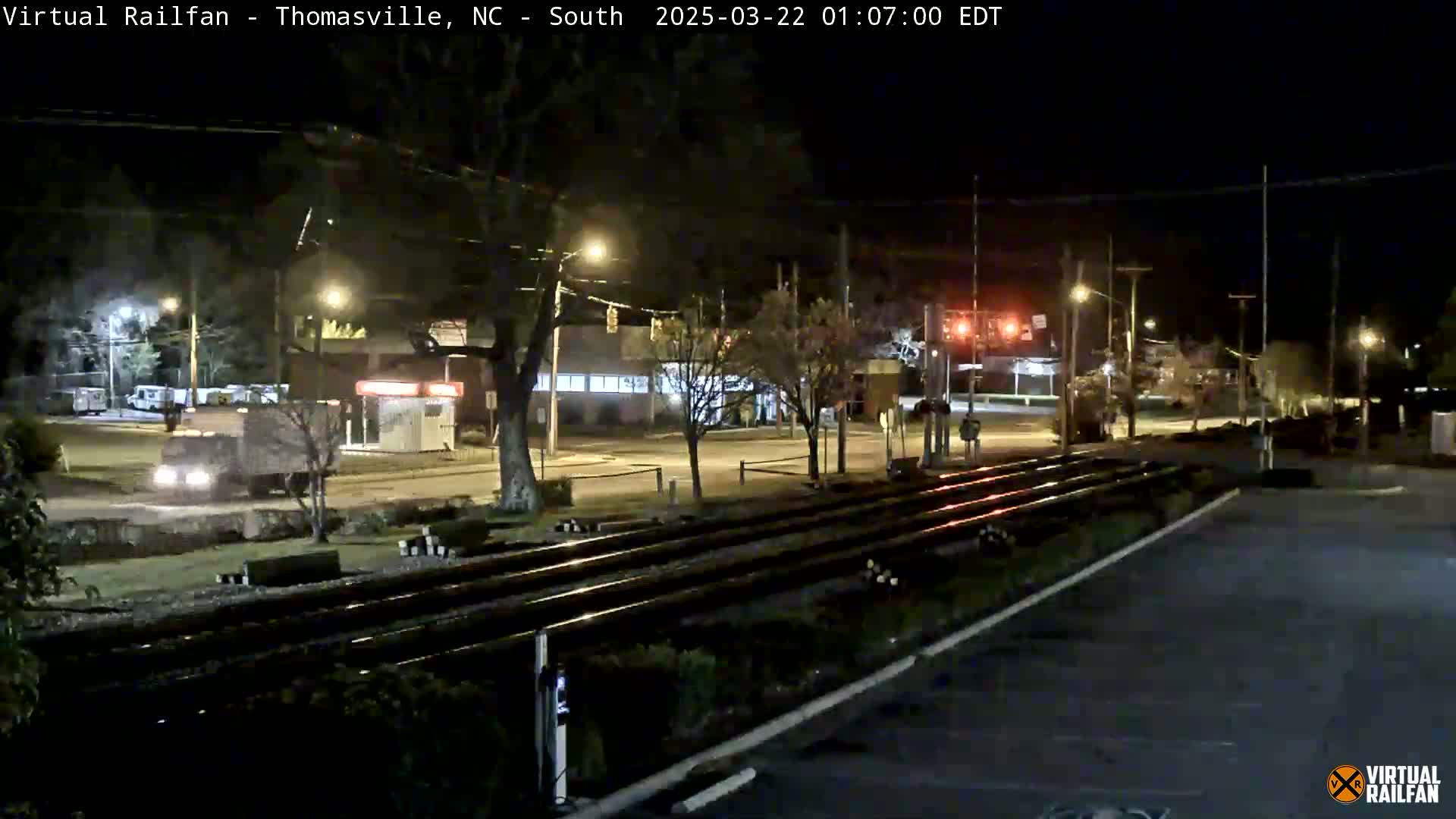 A nighttime view of railroad tracks intersecting a road at a traffic light, with a few vehicles visible under artificial lighting.