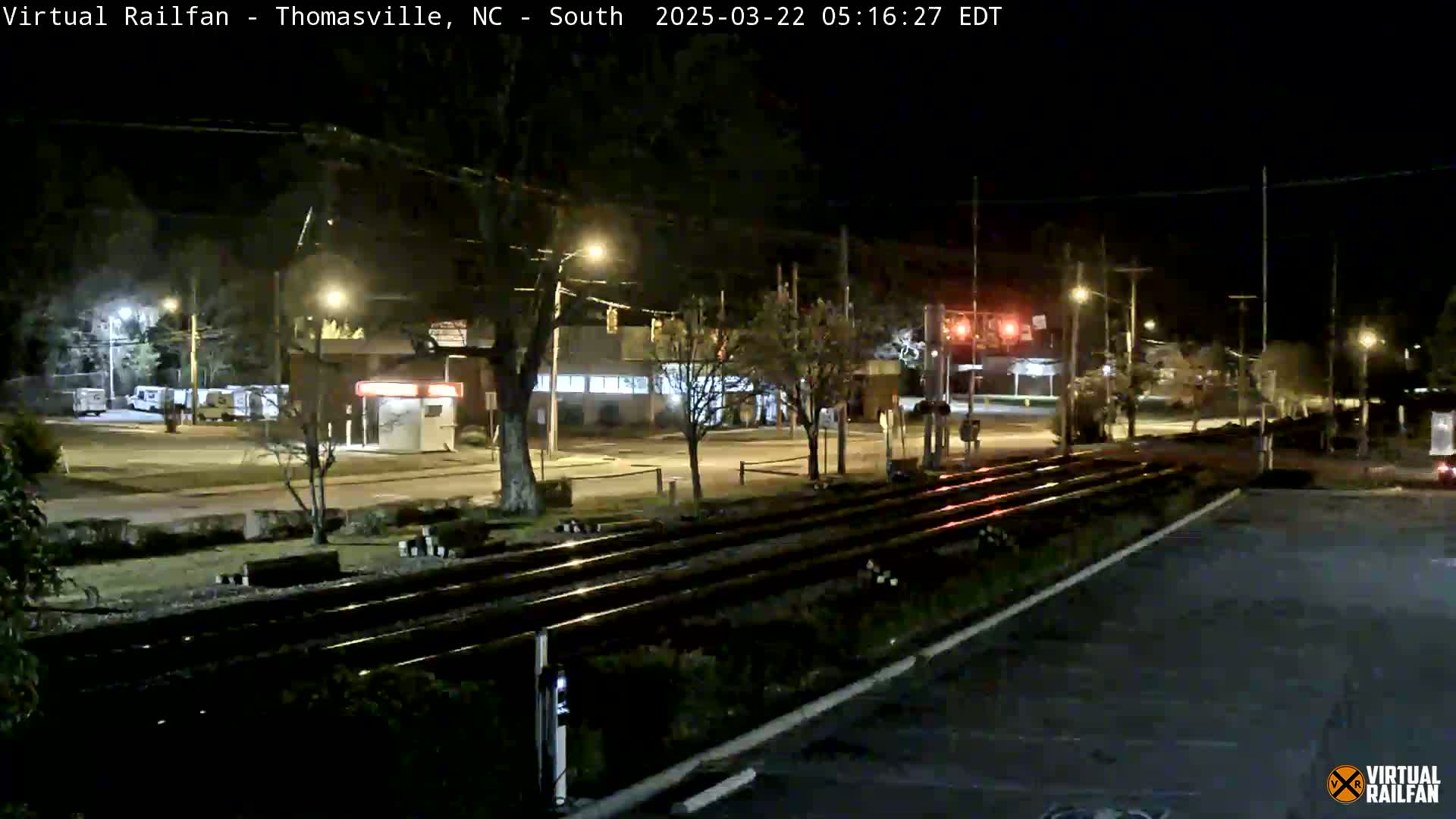 At night, train tracks cross a street in front of buildings and trees under clear skies.