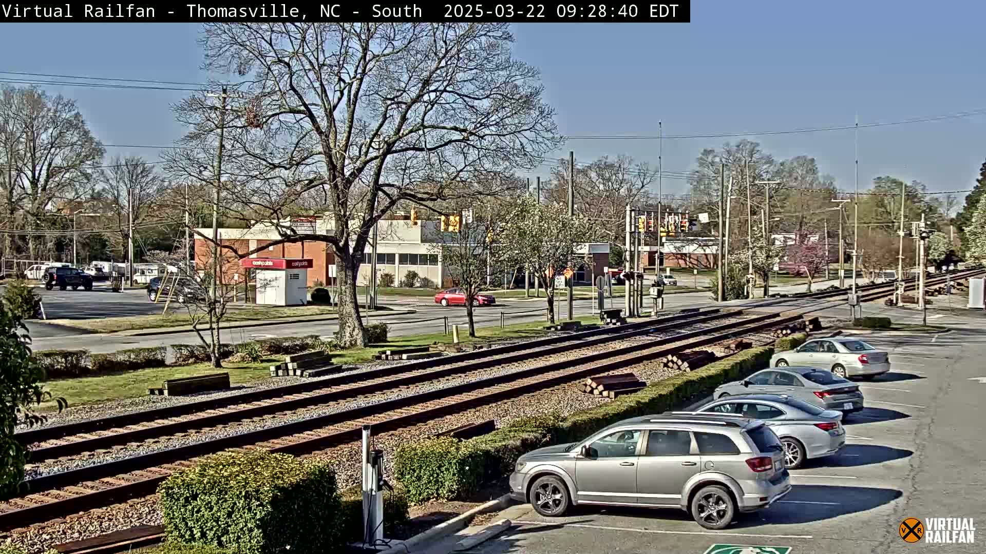 Under a clear blue sky, a view of railroad tracks intersecting with a road shows several cars parked and driving near buildings and trees.