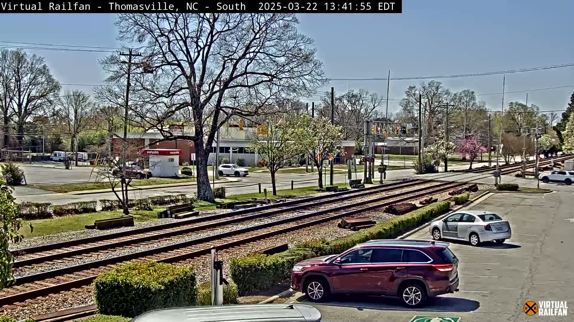 A sunny day view of railroad tracks intersecting a road, with several cars parked nearby and other vehicles on the road, all surrounded by trees and buildings.