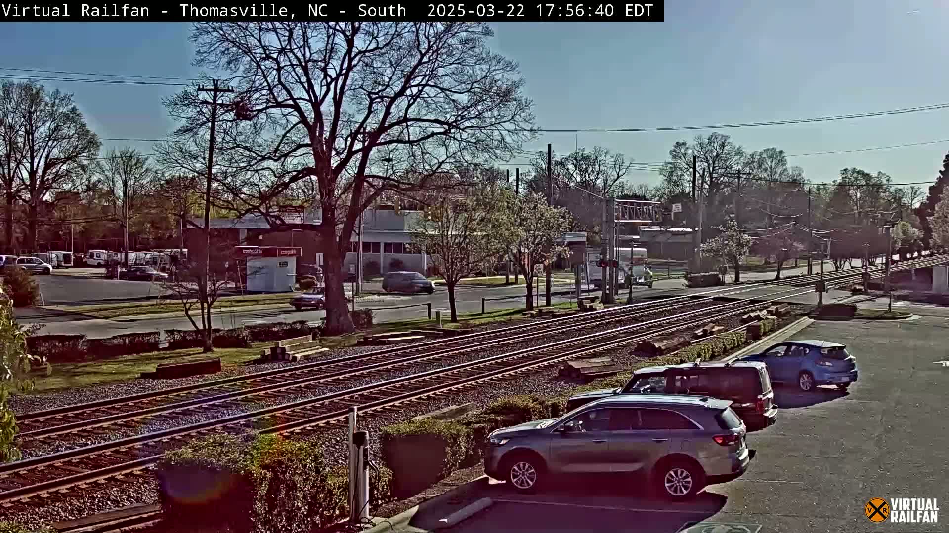 A sunny day view of railroad tracks near a road intersection with several cars parked in a lot and others driving on the road,  trees are bare in the background.