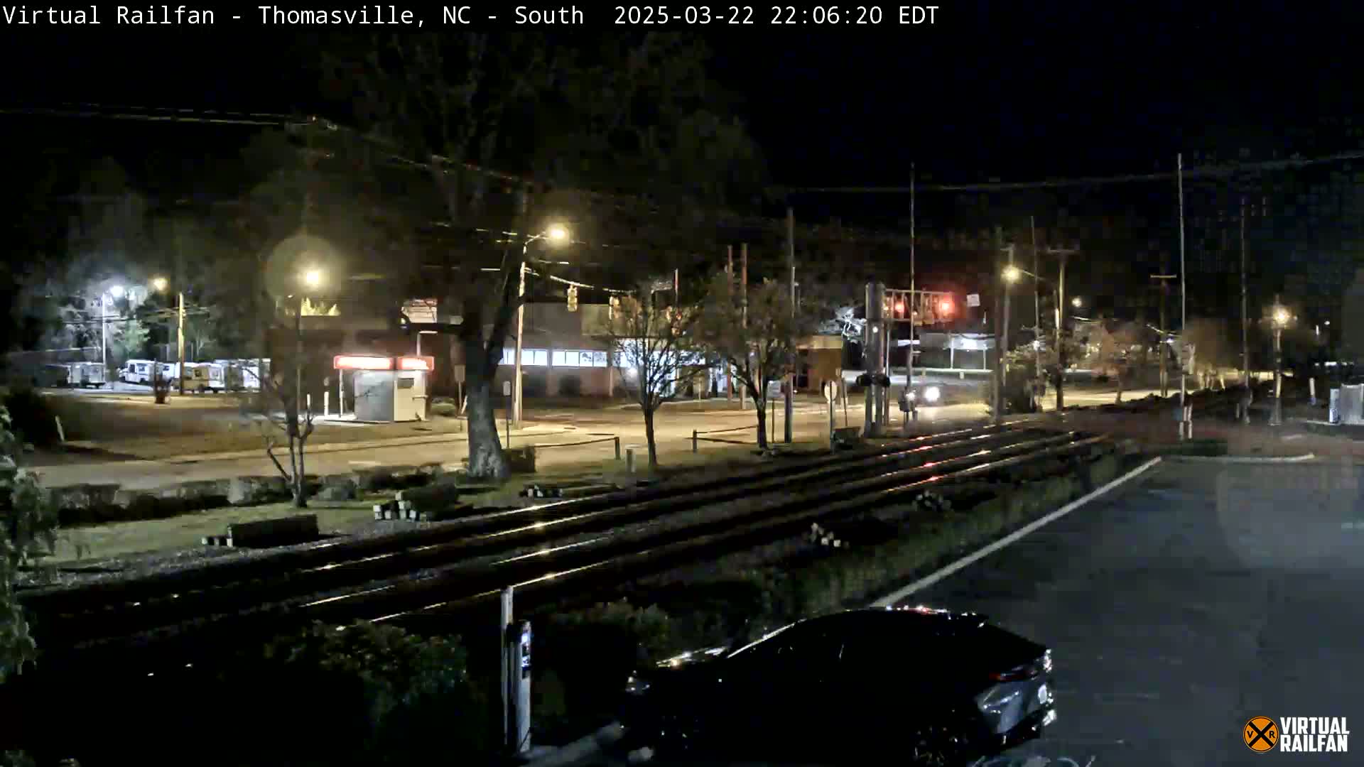 A nighttime view of a railroad crossing near a street intersection with cars and buildings visible under clear skies.