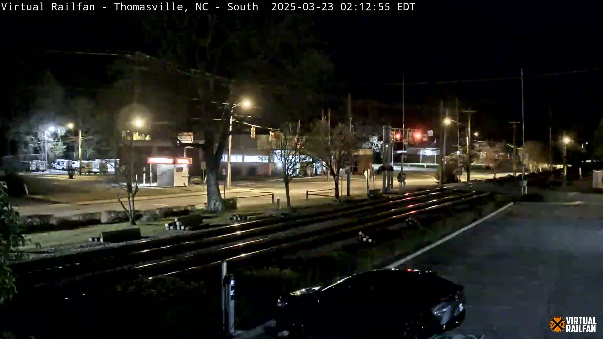 At night, a nighttime view shows train tracks crossing a street, with a few cars visible and streetlights illuminating the scene.