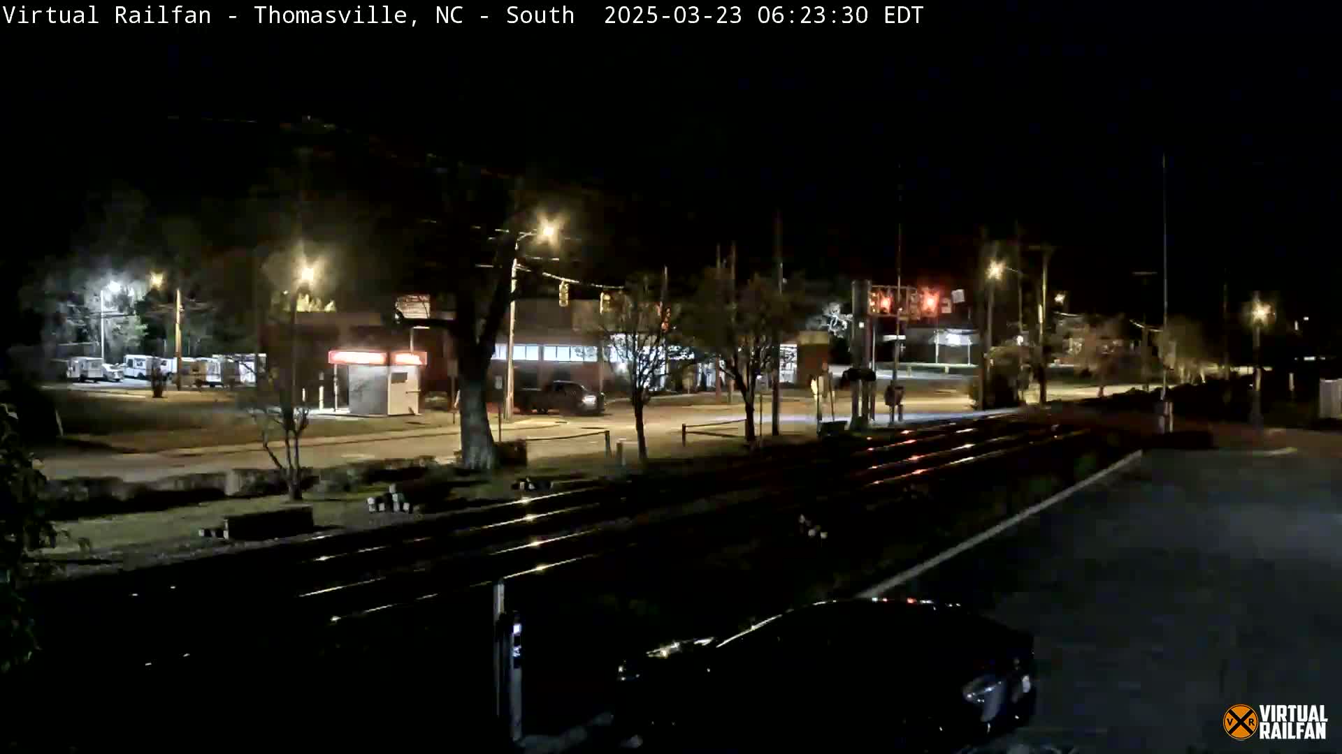 A nighttime view of train tracks crossing a street in front of buildings, under streetlights.