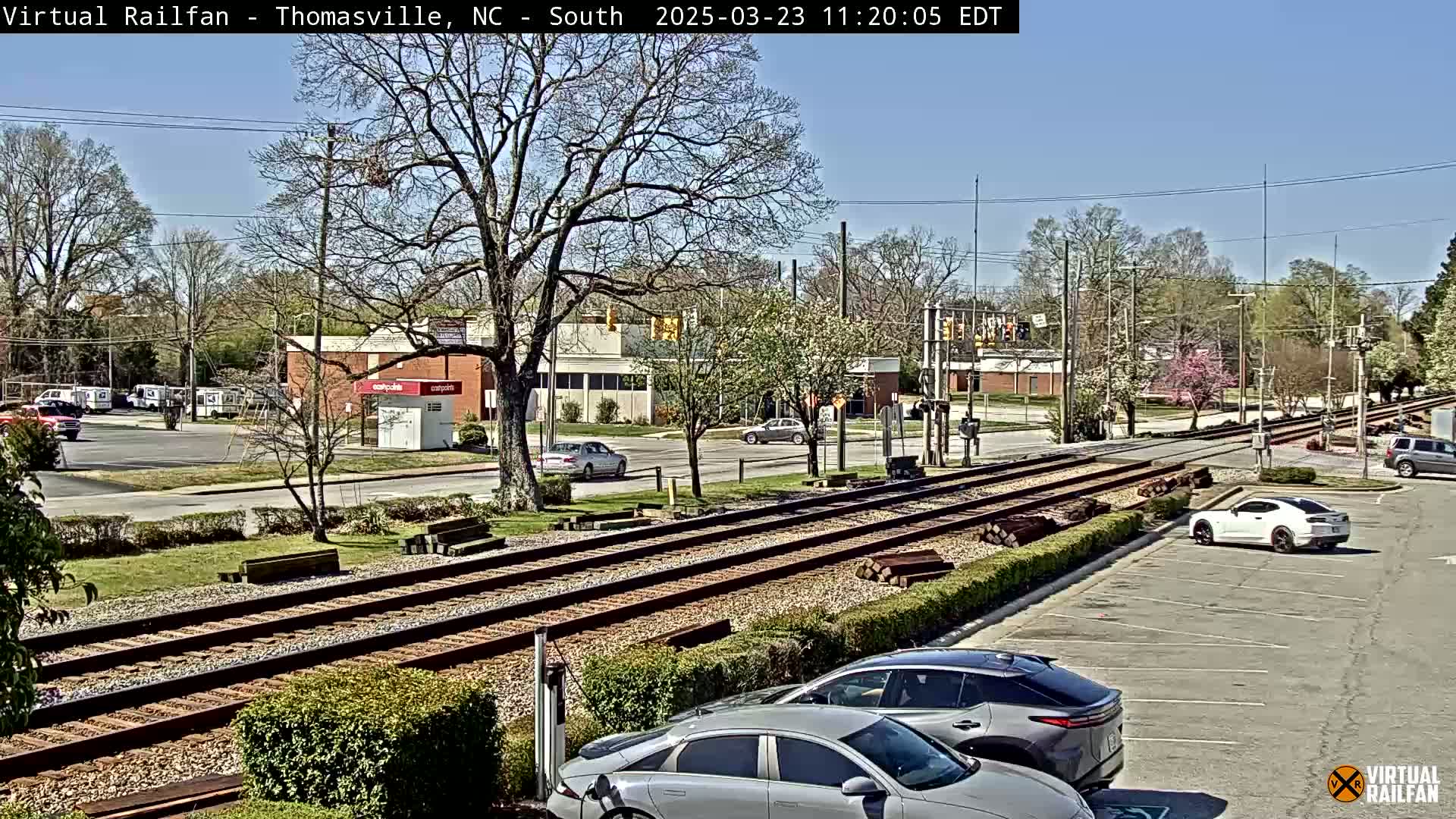 A sunny day view of railroad tracks running through a town, with cars parked in a lot and buildings in the background.