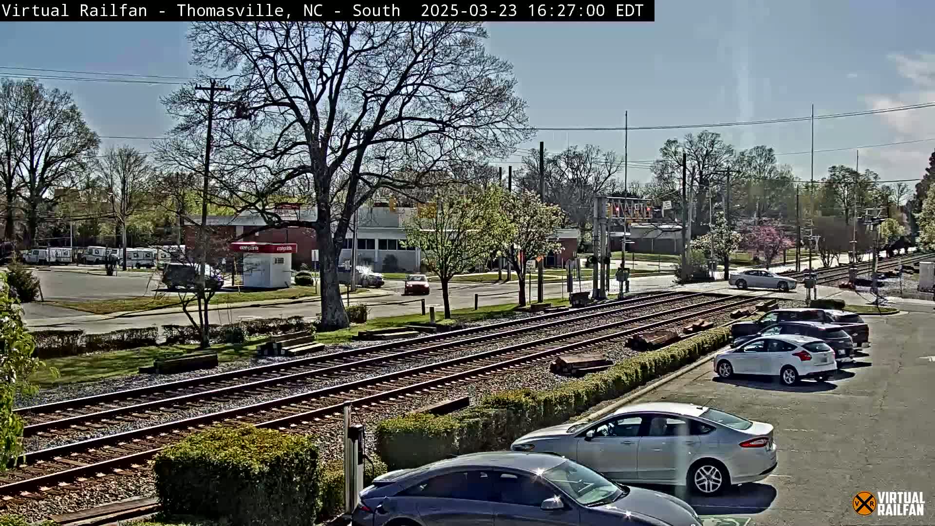 A sunny day shows a street intersection with railroad tracks running through it, several cars parked in a lot nearby, and a few vehicles driving on the road.