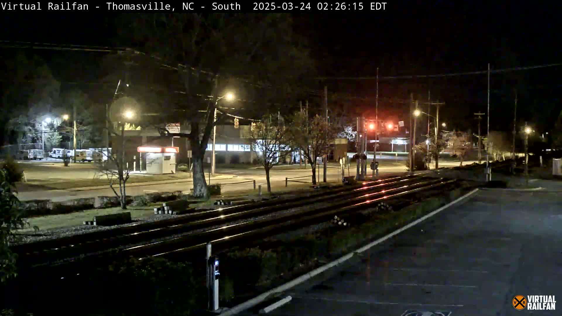 At night, railroad tracks stretch across a mostly empty street scene, passing near buildings and trees under streetlights.