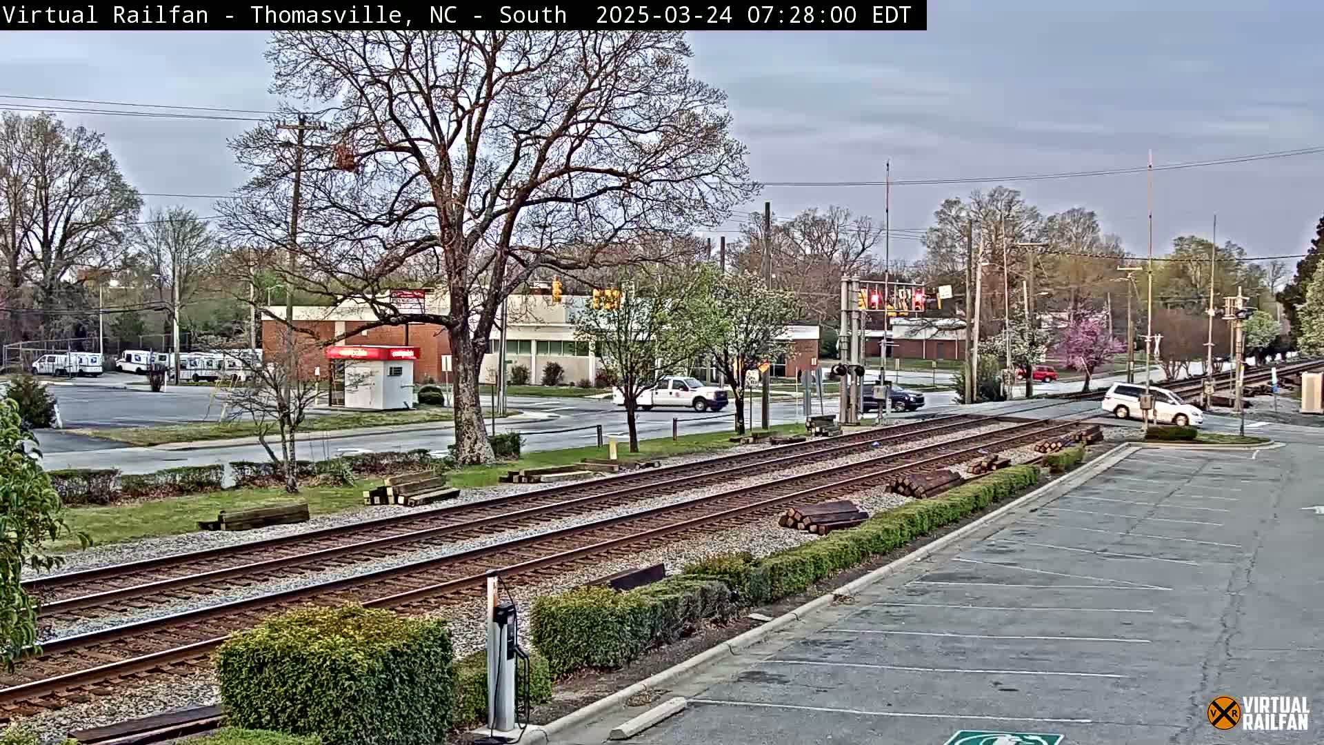 A mostly cloudy day reveals a railroad crossing with several vehicles, including postal trucks, near a commercial building and an empty parking lot.