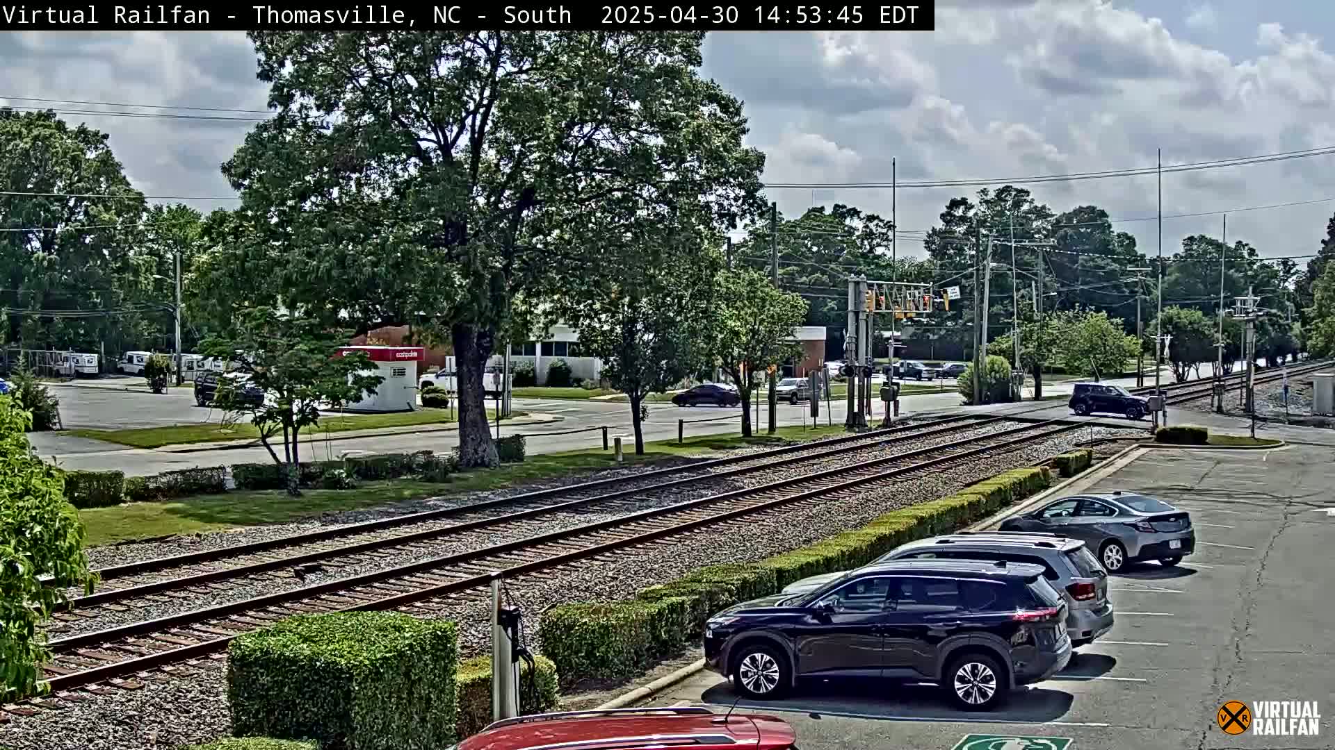 A partly cloudy day shows train tracks intersecting a road with several cars parked nearby and a few cars driving by.