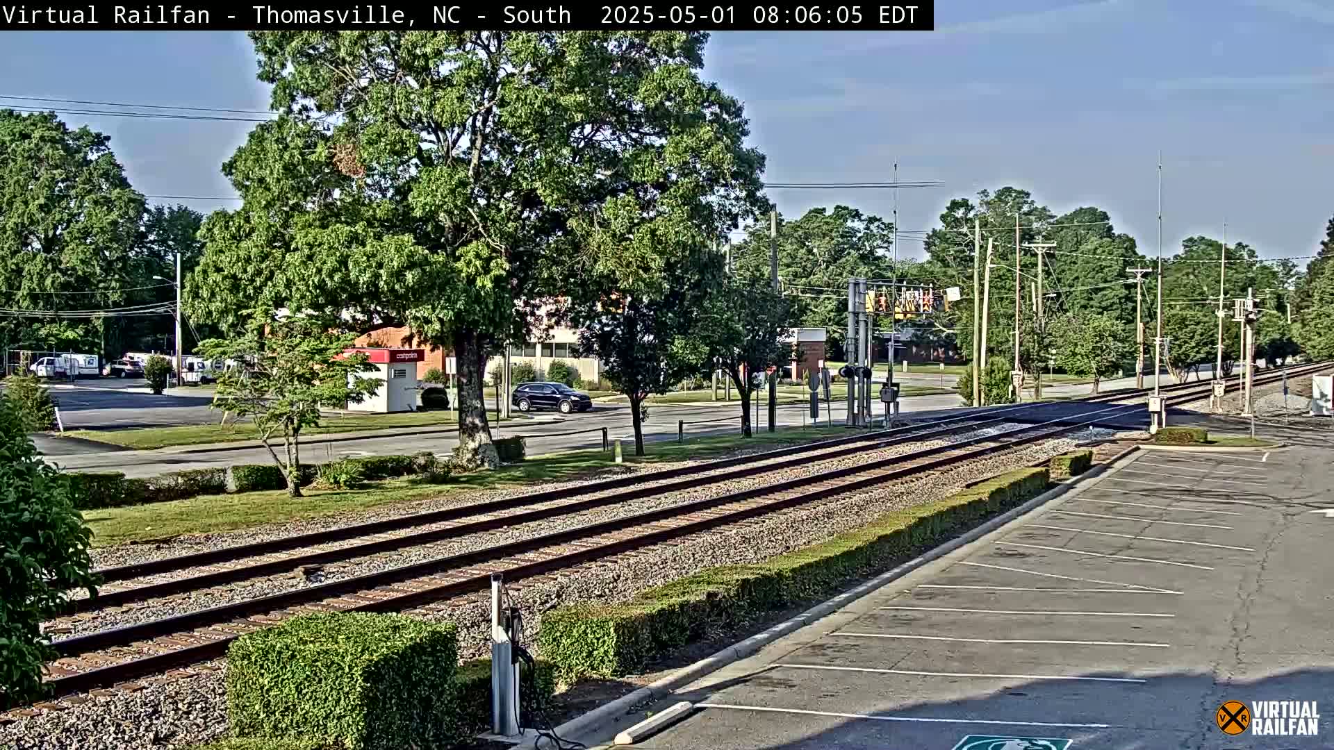 A sunny day view of railroad tracks crossing a road next to a parking lot, with several cars parked and one driving past, surrounded by leafy green trees and bushes.