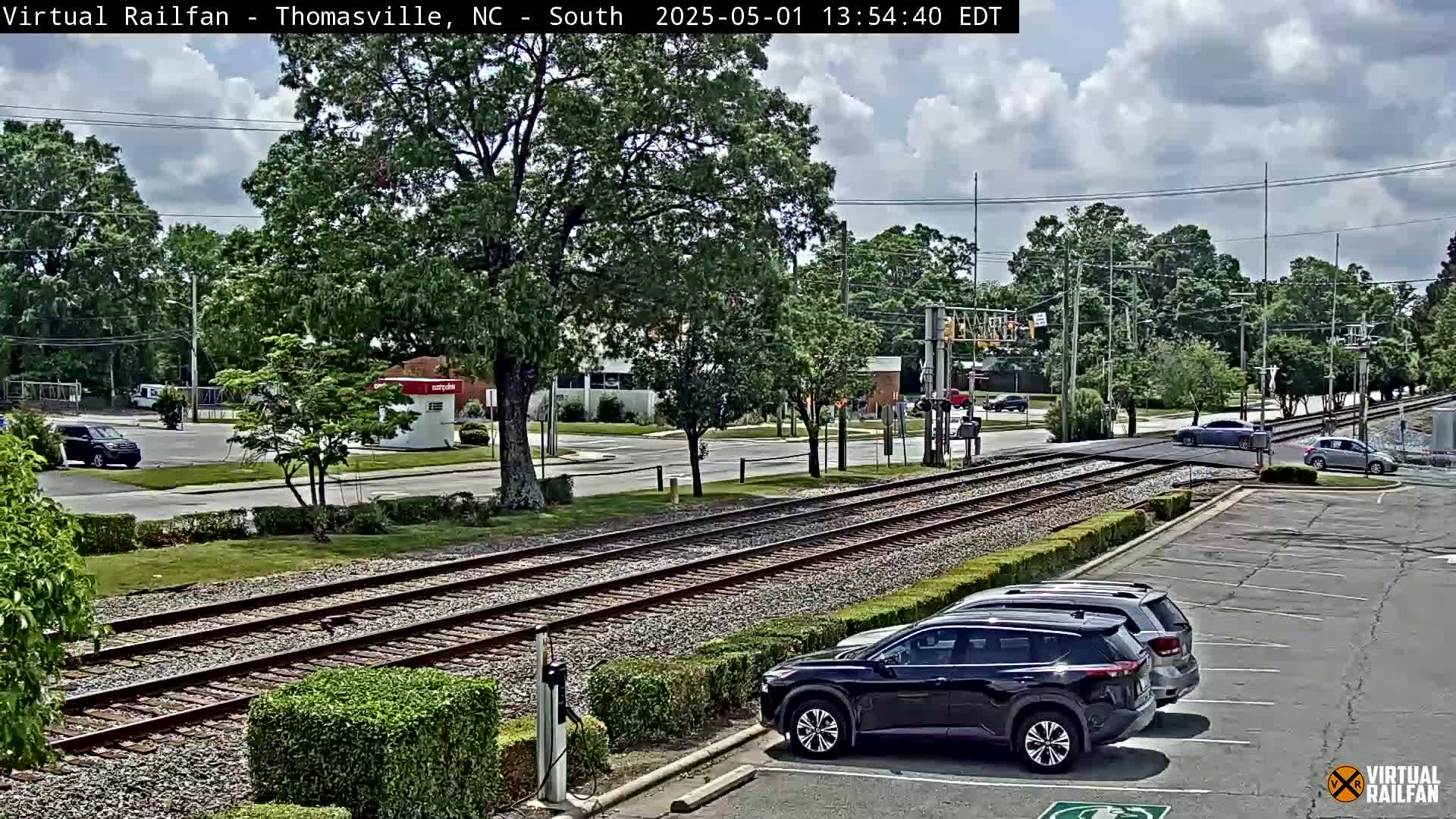 A partly cloudy day shows a railroad crossing near a street intersection with several cars parked in a lot and some driving on the road.