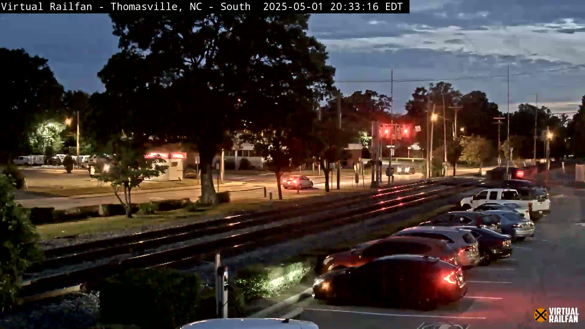 A twilight scene shows a railroad crossing with cars parked in a lot and a few cars driving on the road, under a partly cloudy sky.