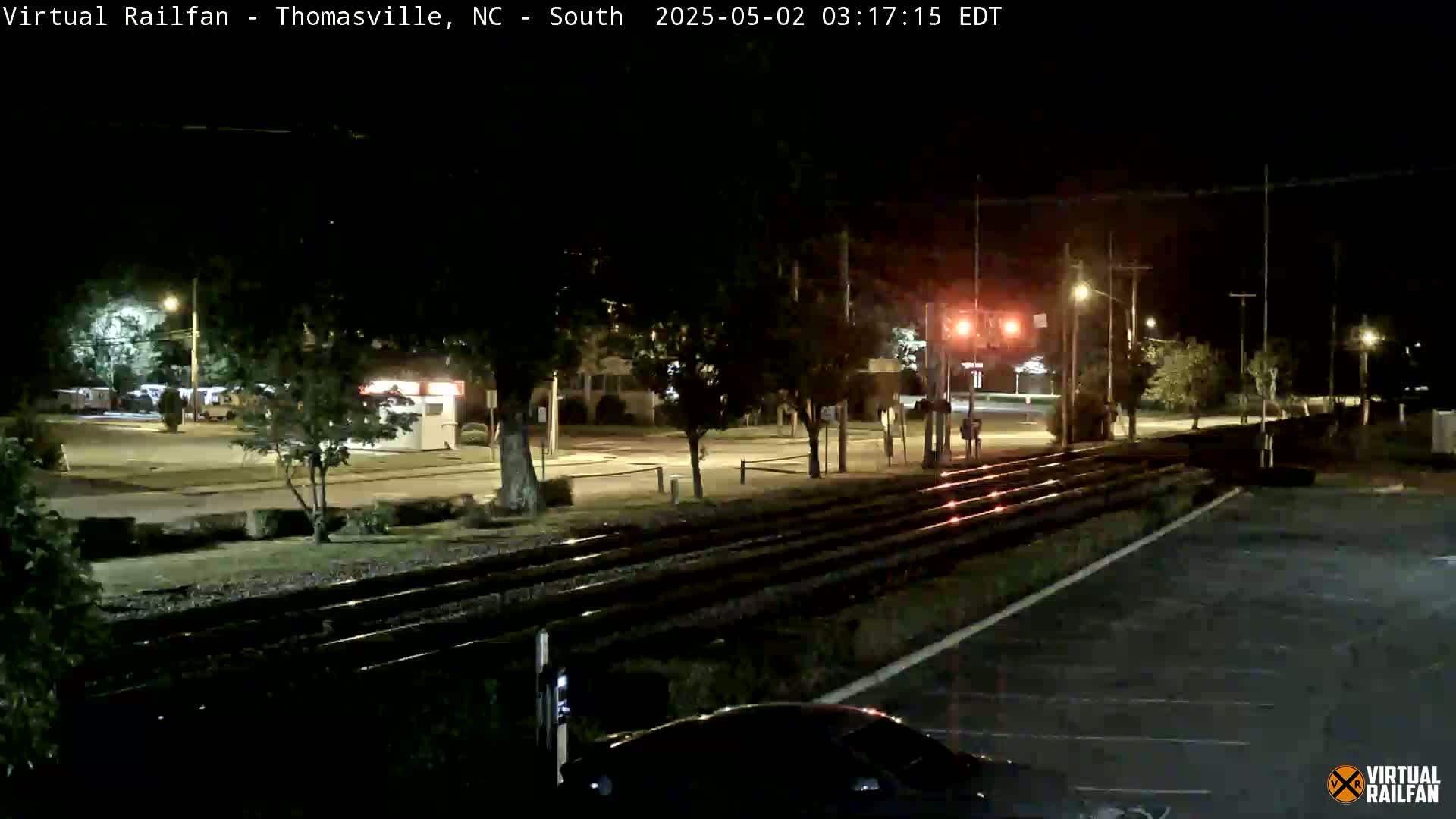 At night, railroad tracks intersect a street with sparse nighttime traffic under streetlights, and a few cars are visible in the parking lot near the camera.