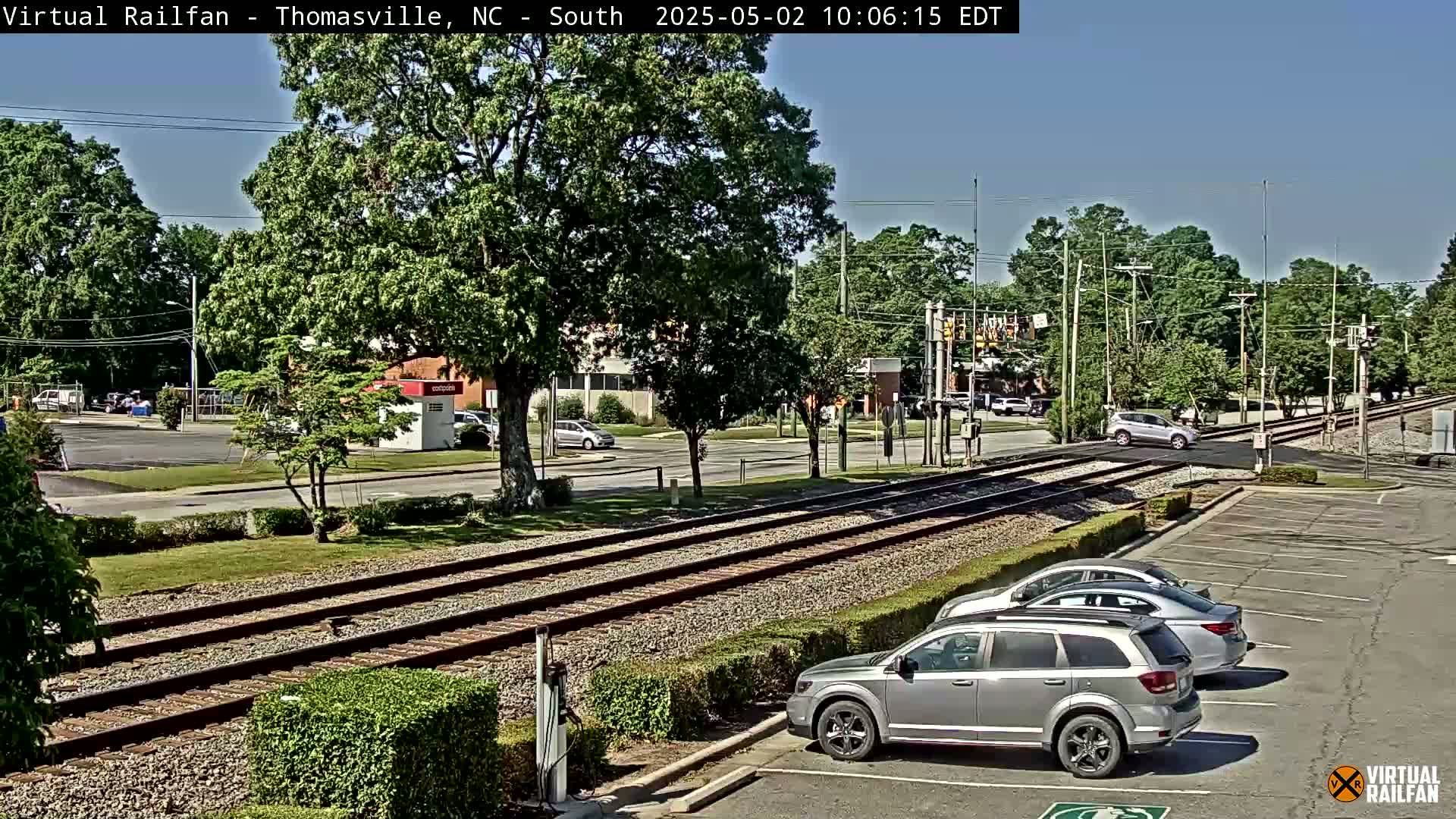 A sunny day view of railroad tracks crossing a road, with several cars parked in a lot nearby and more vehicles visible on the road.