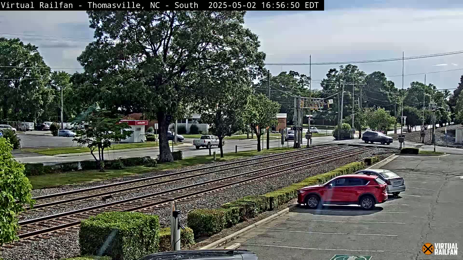 A partly sunny day shows train tracks crossing a road with several cars parked and driving near lush green landscaping.