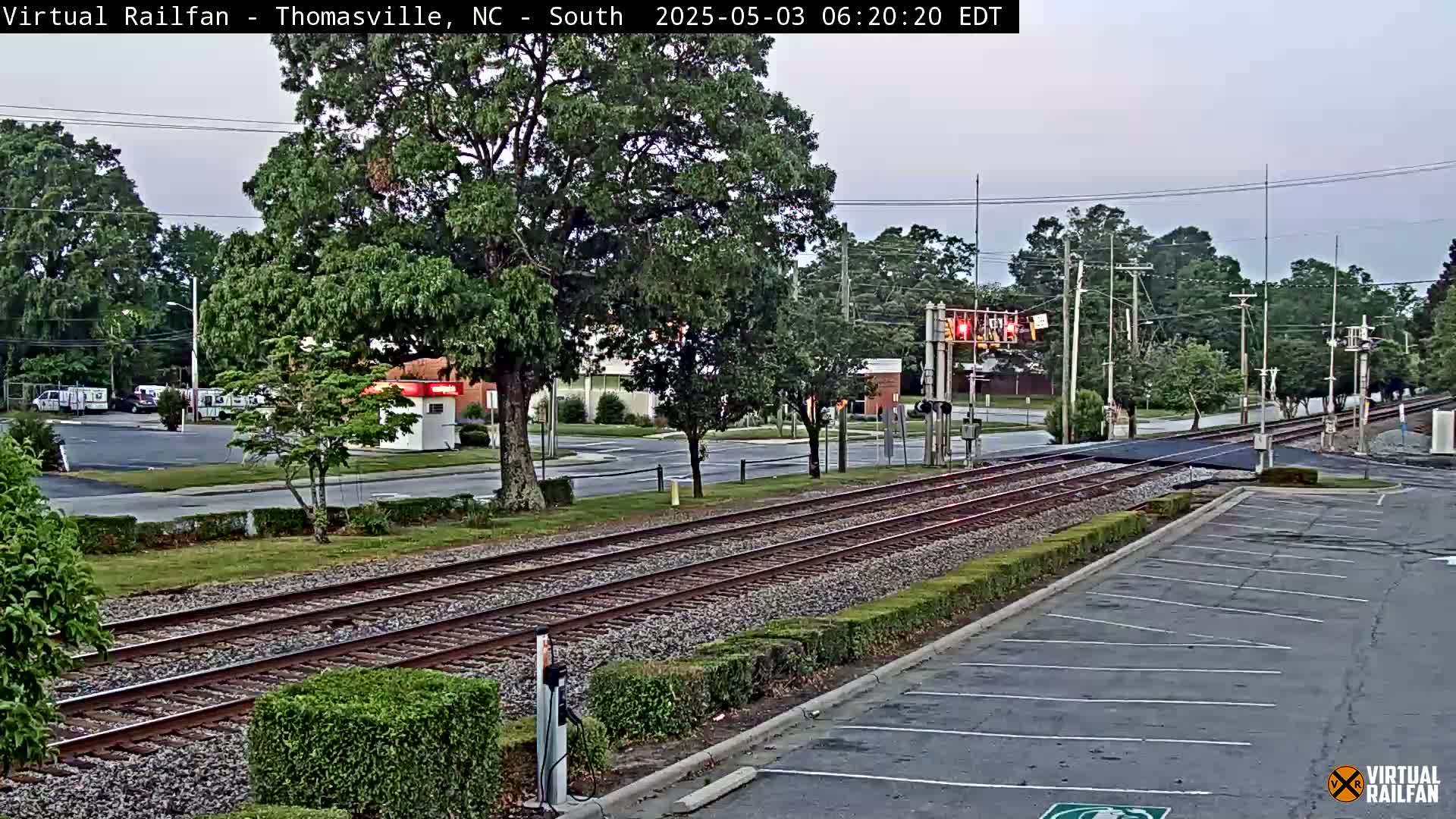 Train tracks intersect a road under a mostly clear, twilight sky, with trees and buildings in the background.
