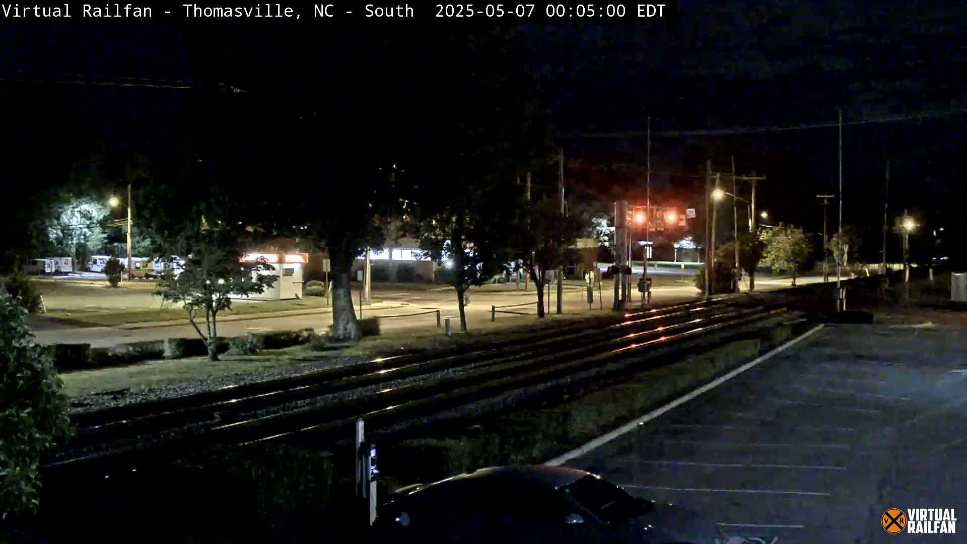 A nighttime view of railroad tracks next to a road and parking lot, with trees and buildings visible in the background under a clear, dark sky.