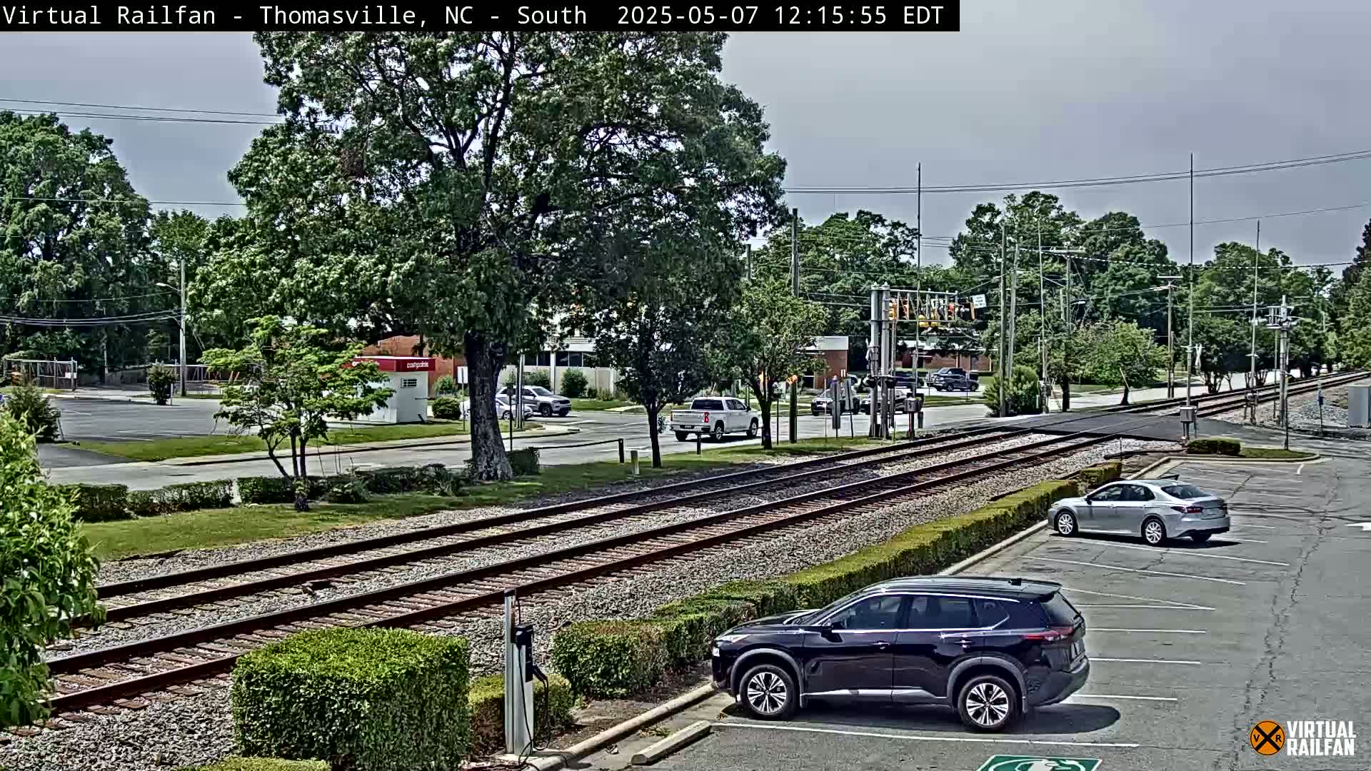 Under a partly cloudy sky, a view of railroad tracks intersecting a road shows several cars parked and driving near a railway crossing, with trees and buildings in the background.