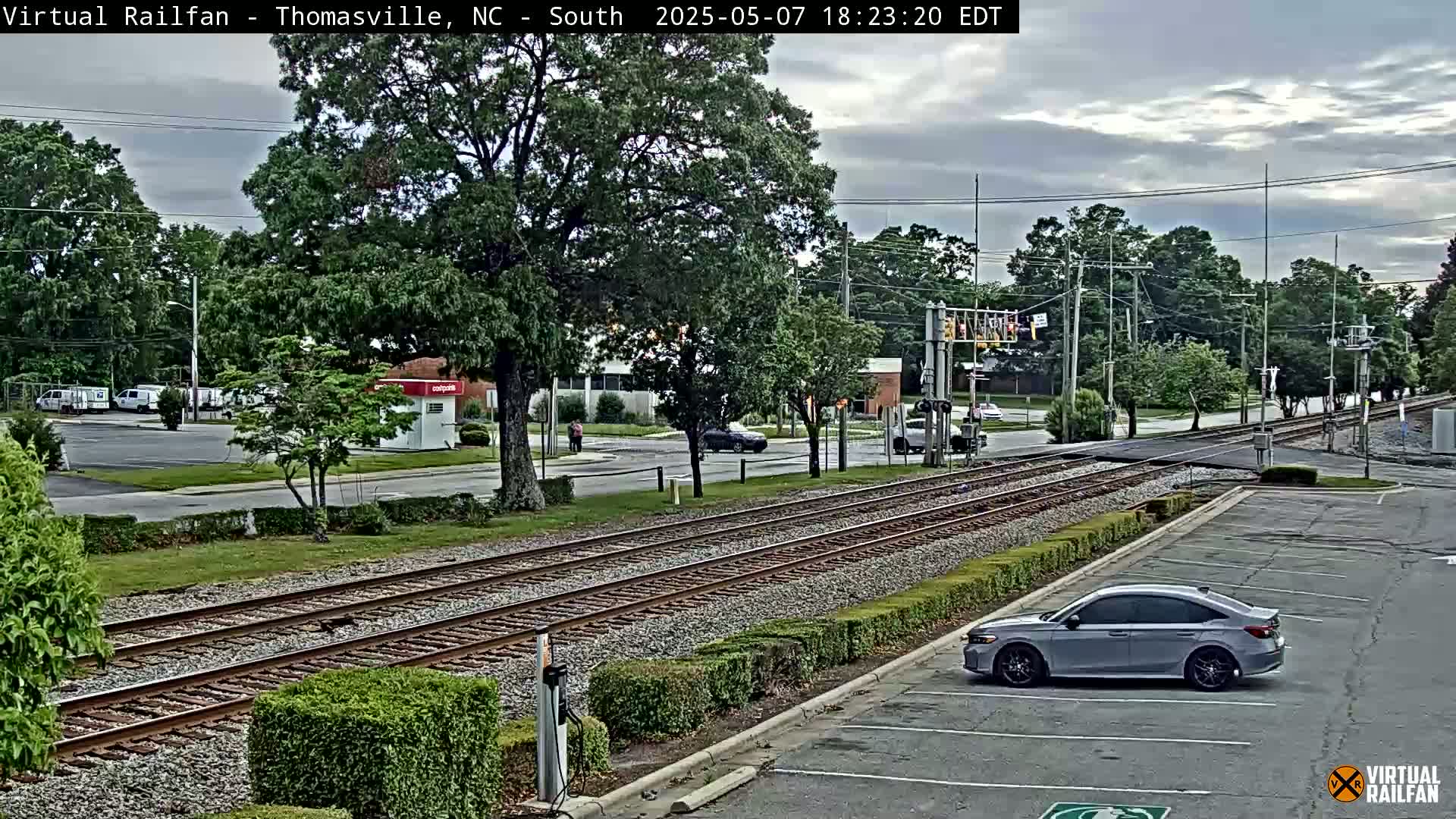A mostly cloudy day shows a silver car parked in a lot next to railroad tracks intersecting a road with several other vehicles visible.