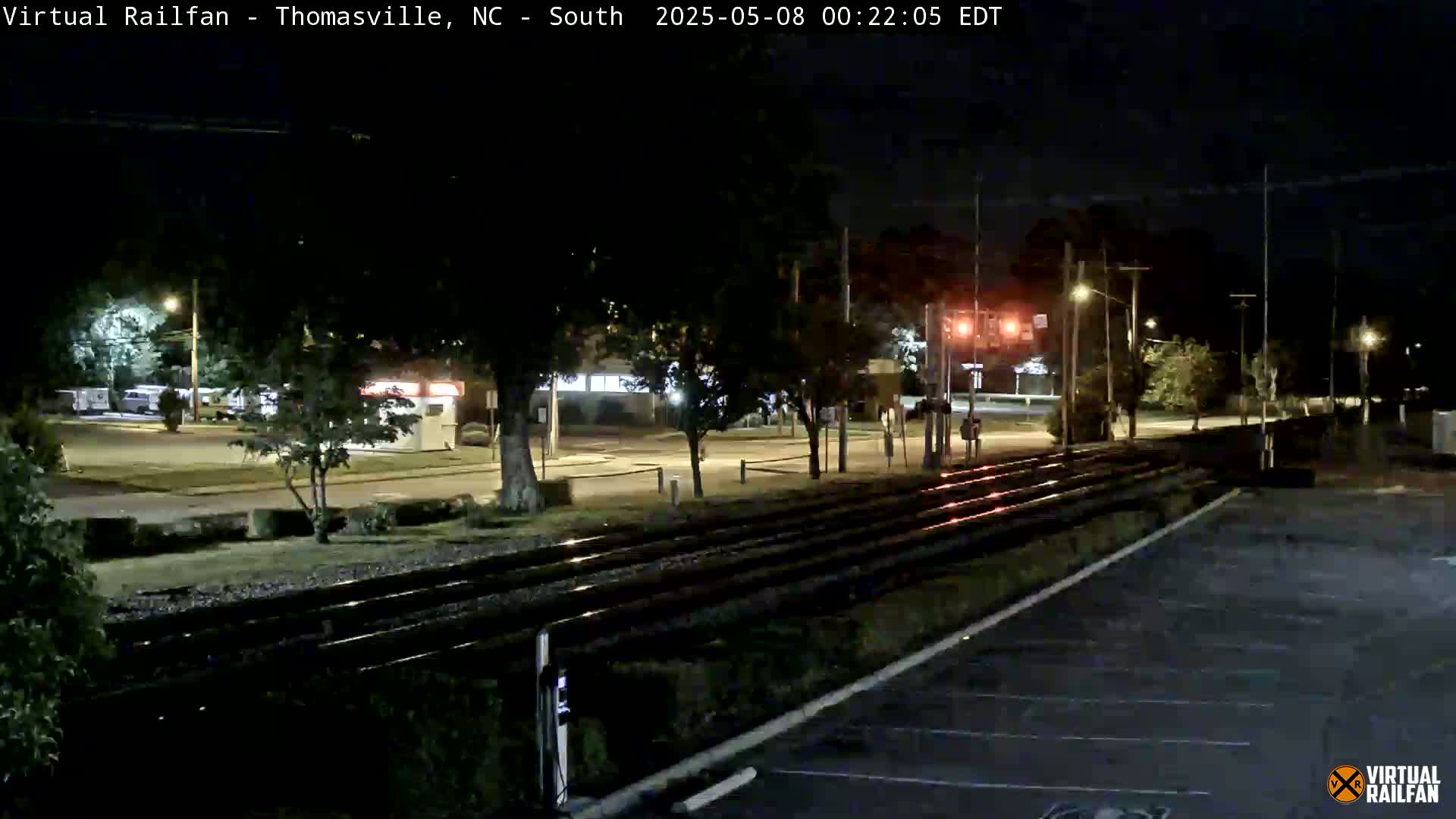 At night, a view of train tracks runs alongside a mostly empty parking lot, bordered by trees and streetlights illuminating a road and buildings in the distance.