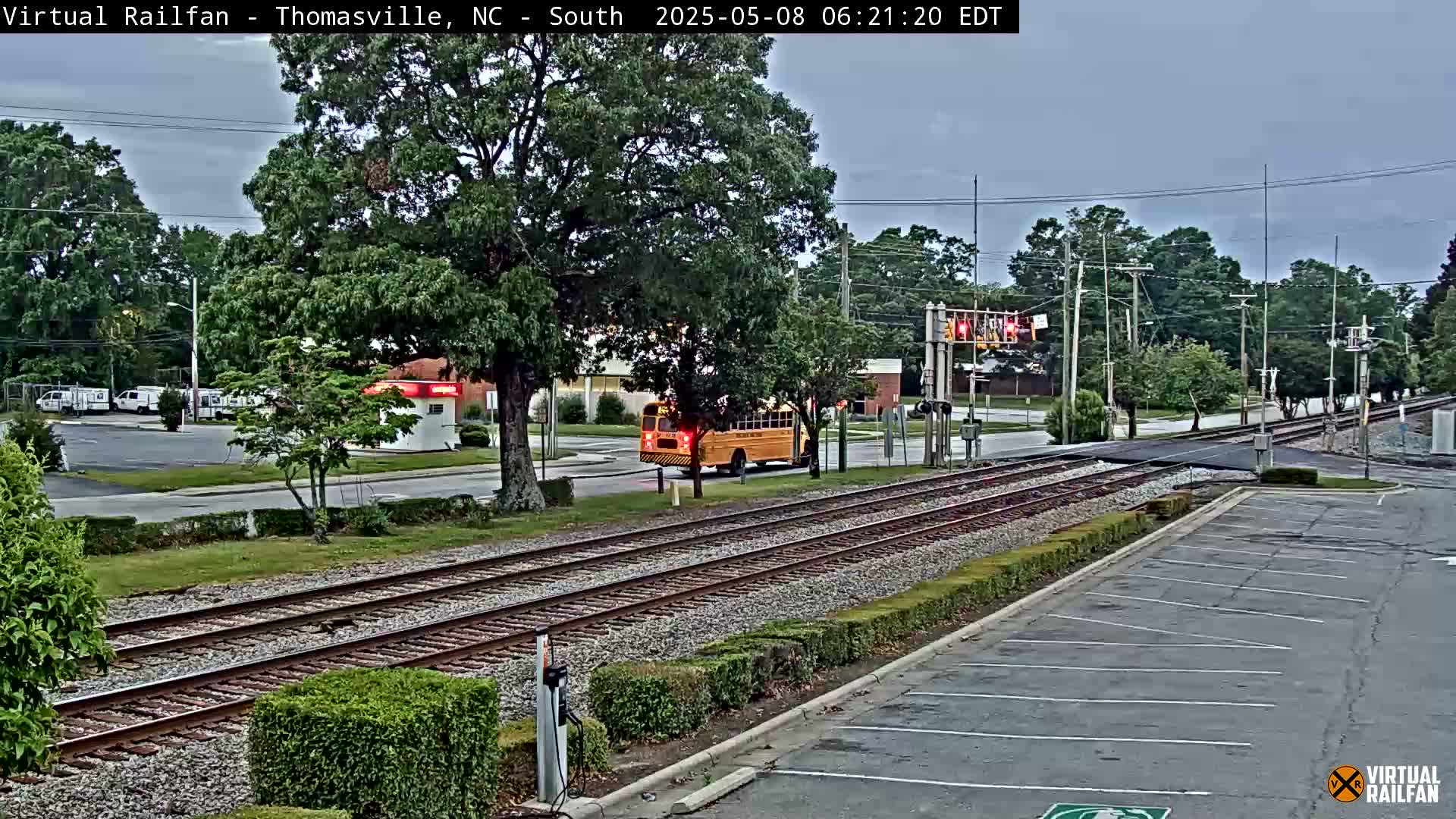 A school bus is stopped at a railroad crossing on a partly cloudy day, with train tracks visible in the foreground and a parking lot in the background.