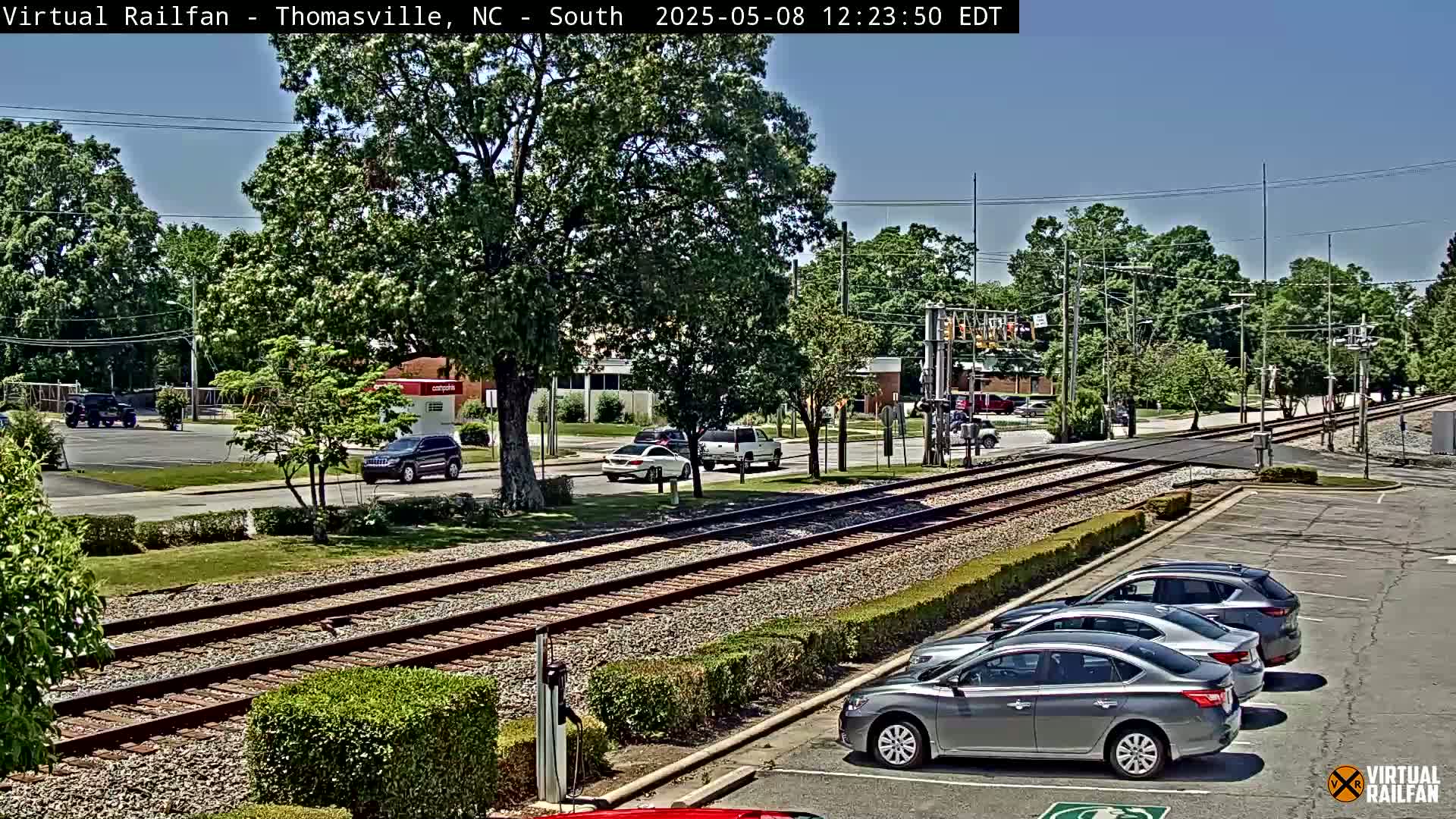 A sunny day view of a railroad crossing with several cars parked in a lot and a few driving on the road, surrounded by green trees and bushes.