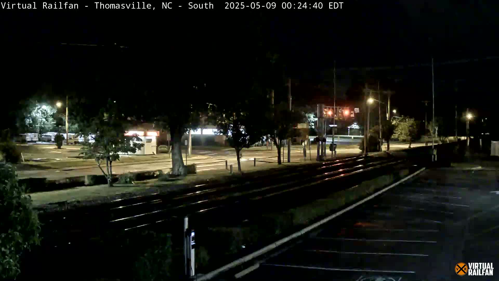 A nighttime view of train tracks running through a mostly empty parking lot and a street, with sparse streetlights illuminating the scene.