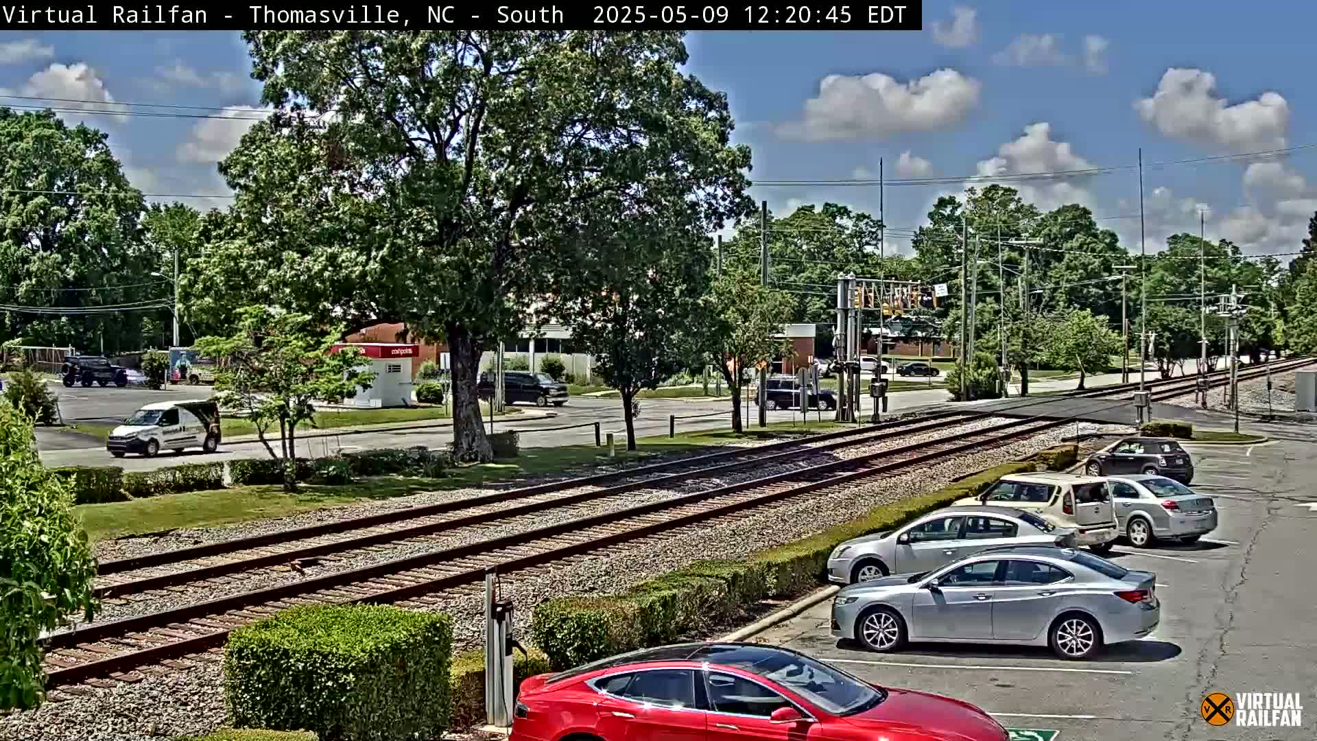 A partly sunny view of a railroad crossing with several cars parked in a lot next to it, and other cars driving on the road.