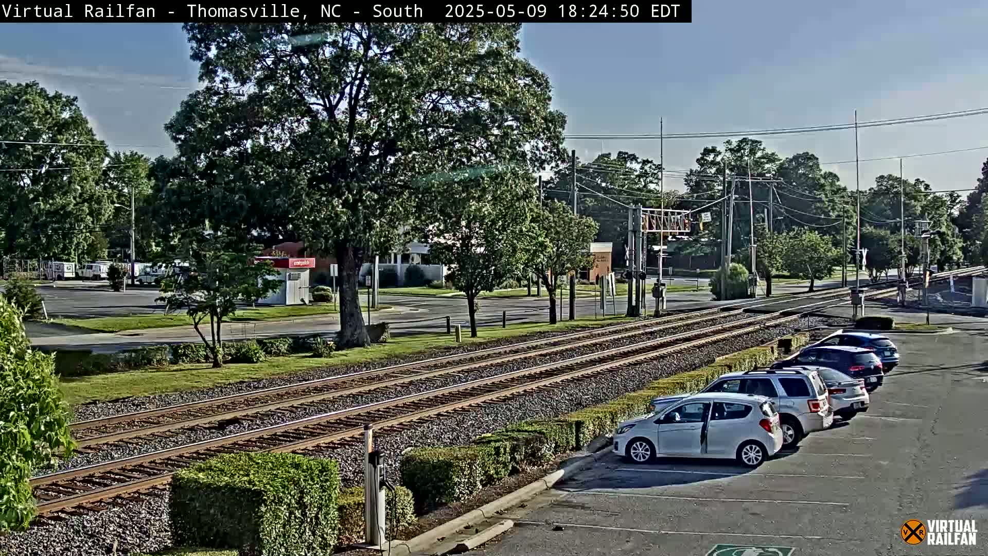 A sunny view of train tracks running through a town, with several cars parked in a lot next to the tracks and lush green trees and landscaping surrounding the area.