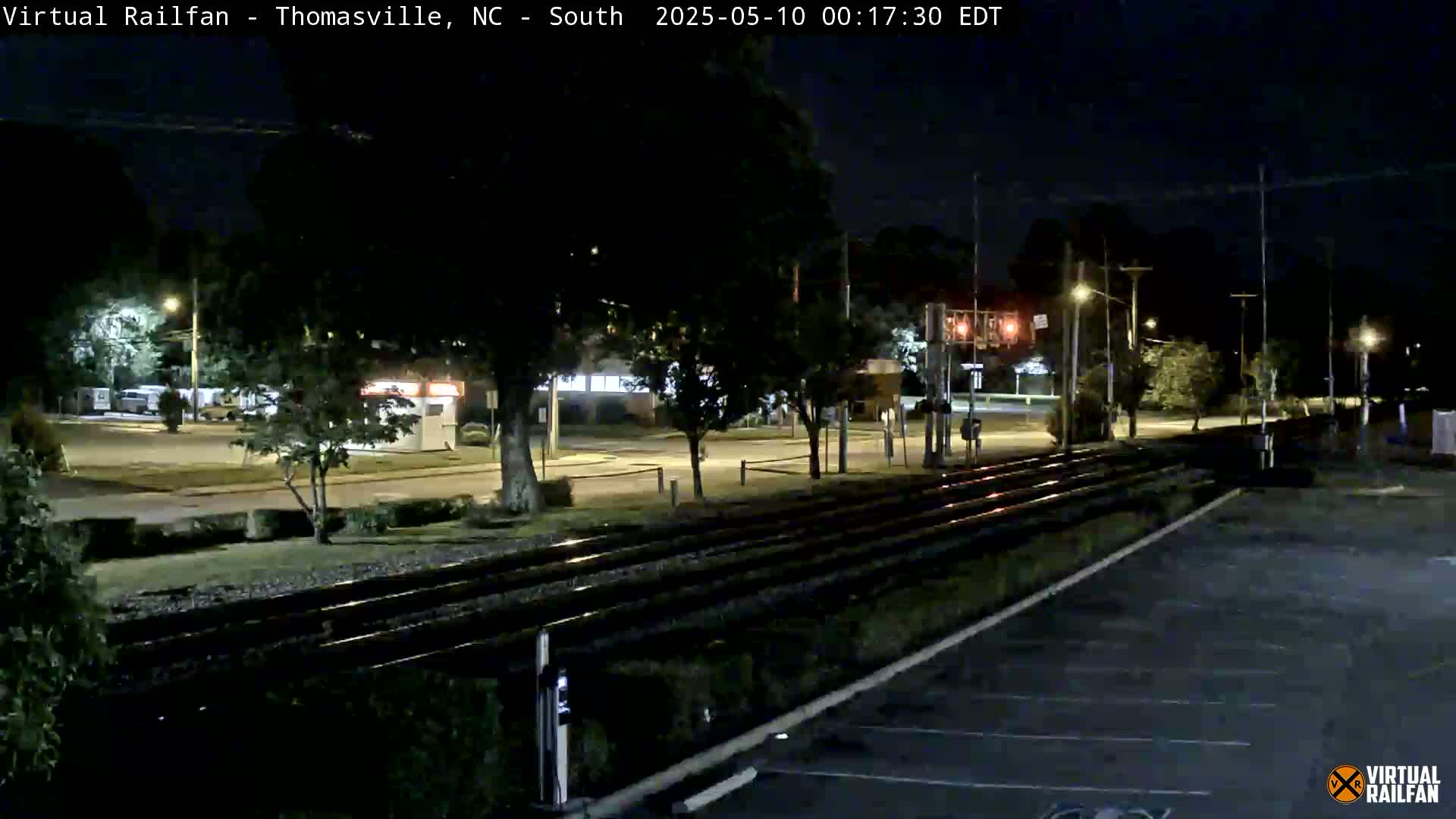 At night, train tracks run alongside a mostly empty parking lot next to a road with sparsely lit buildings and trees.