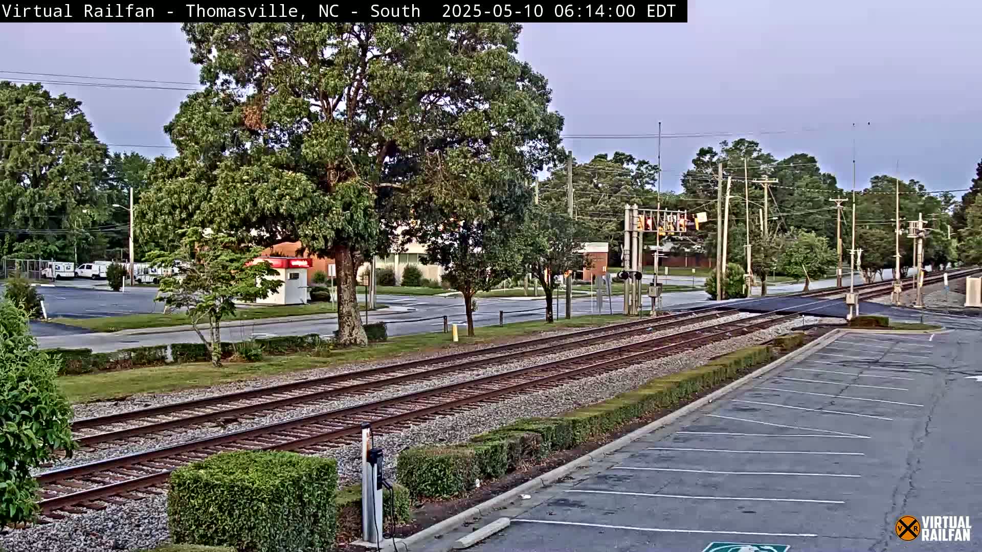 Under a partly cloudy sky, railroad tracks cross a road next to a parking lot and several trees.