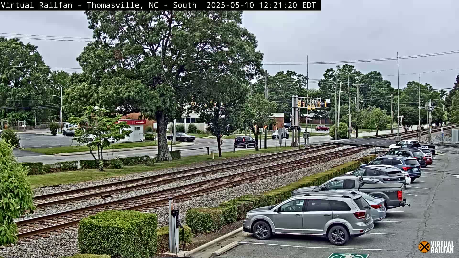 A view of railroad tracks running through a town on an overcast day, with several cars parked in a lot beside them and other cars driving on a nearby road.