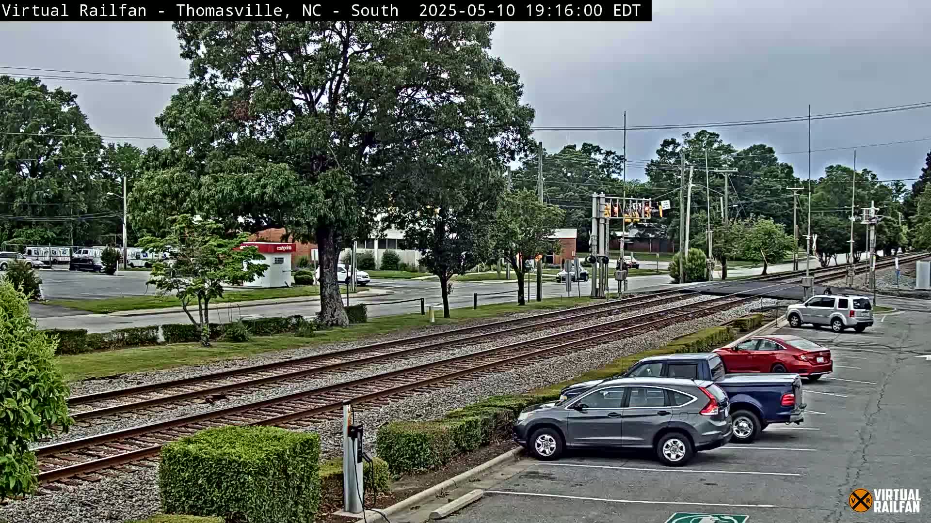 A view of railroad tracks intersecting a road, with several cars parked nearby under an overcast sky.