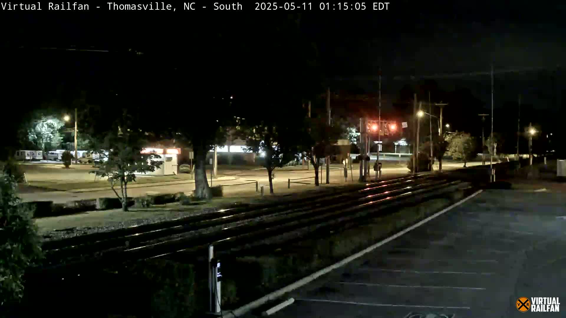 At night, train tracks run through a mostly empty parking lot and street, near buildings and trees under a clear sky.