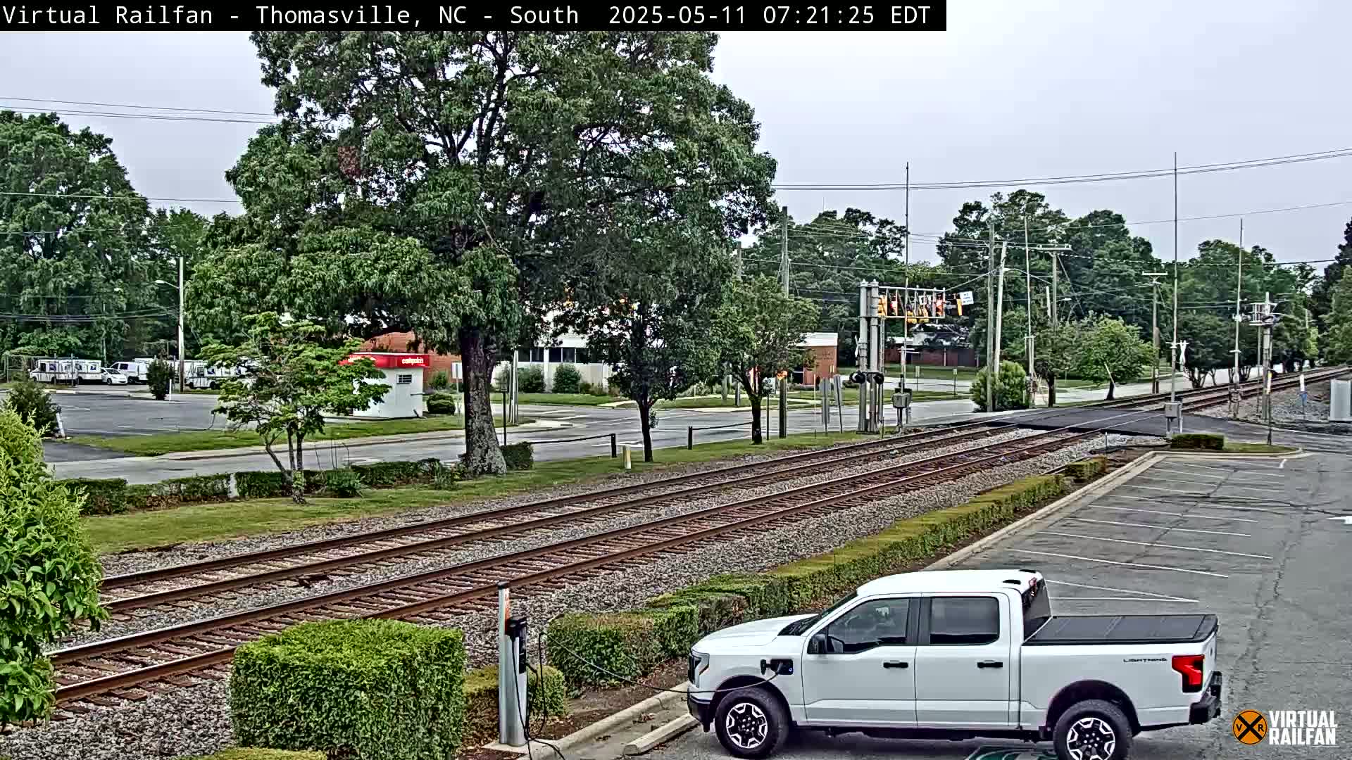 A white pickup truck is parked near railroad tracks that cross a road, under an overcast sky.