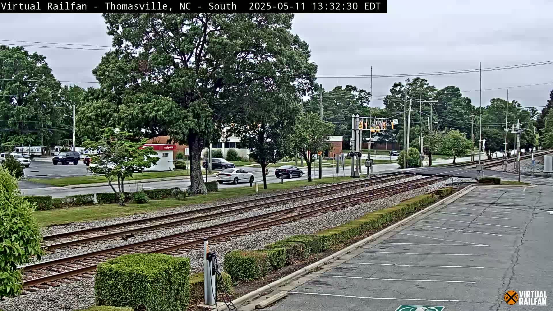 A view of railroad tracks intersecting a road, with several cars visible on the road and trees lining the street under an overcast sky.