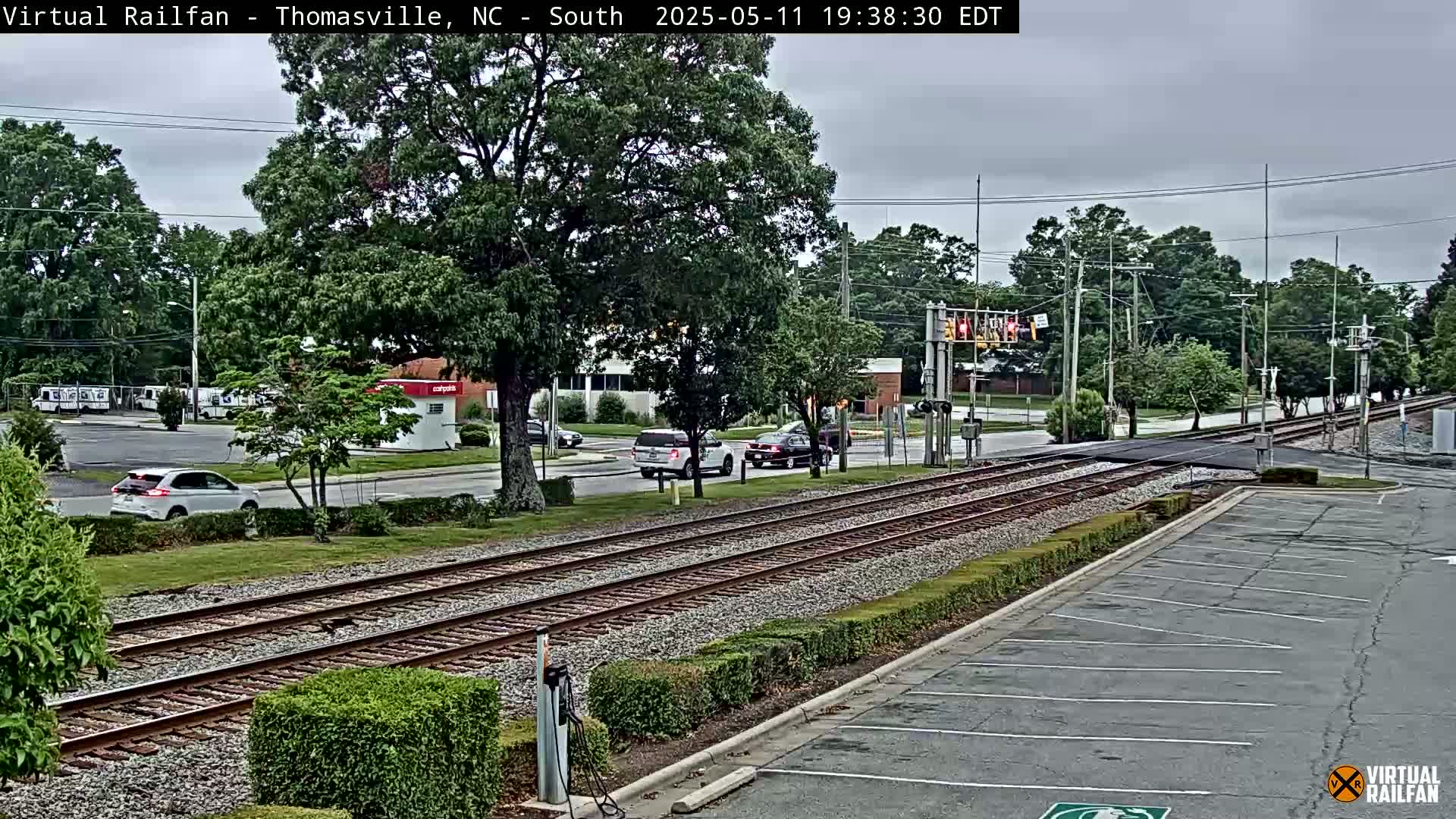 A cloudy day shows a railroad crossing with several vehicles at an intersection, including a white SUV and a silver car, near a parking lot and some green shrubbery.